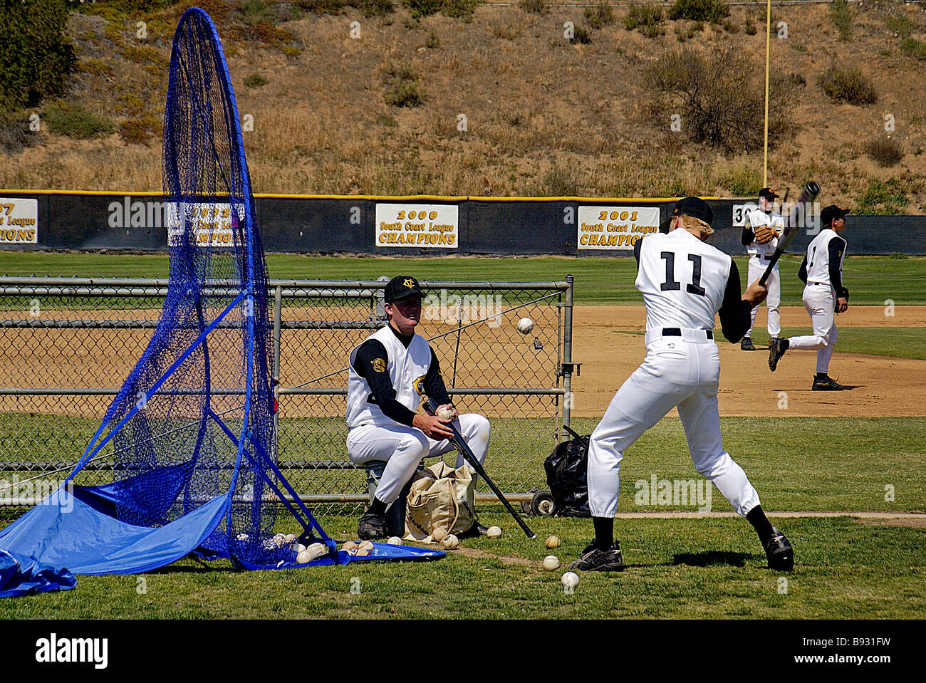Während eine sitzende Teamkollege Kugeln wirft ein Baseball Spieler Praktiken mit der Wimper in ein blaues Sicherheitsnetz am California High school Stockfoto