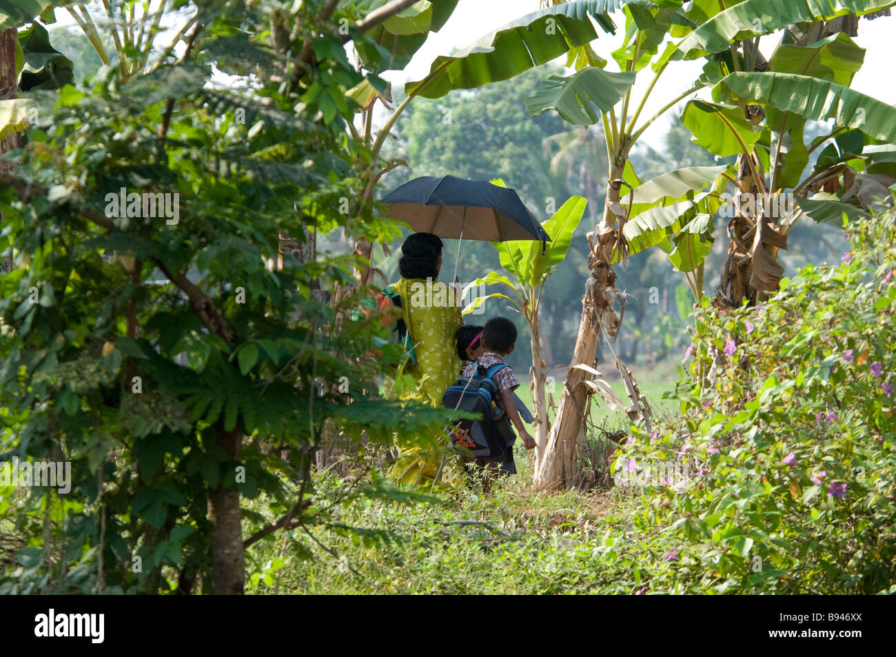 Kinder gehen von der Schule nach Hause mit Mutter holding Regenschirm als Sonnenschirm in die Landschaft am Ufer eines Flusses auf die Kerala Backwaters, Indien Stockfoto