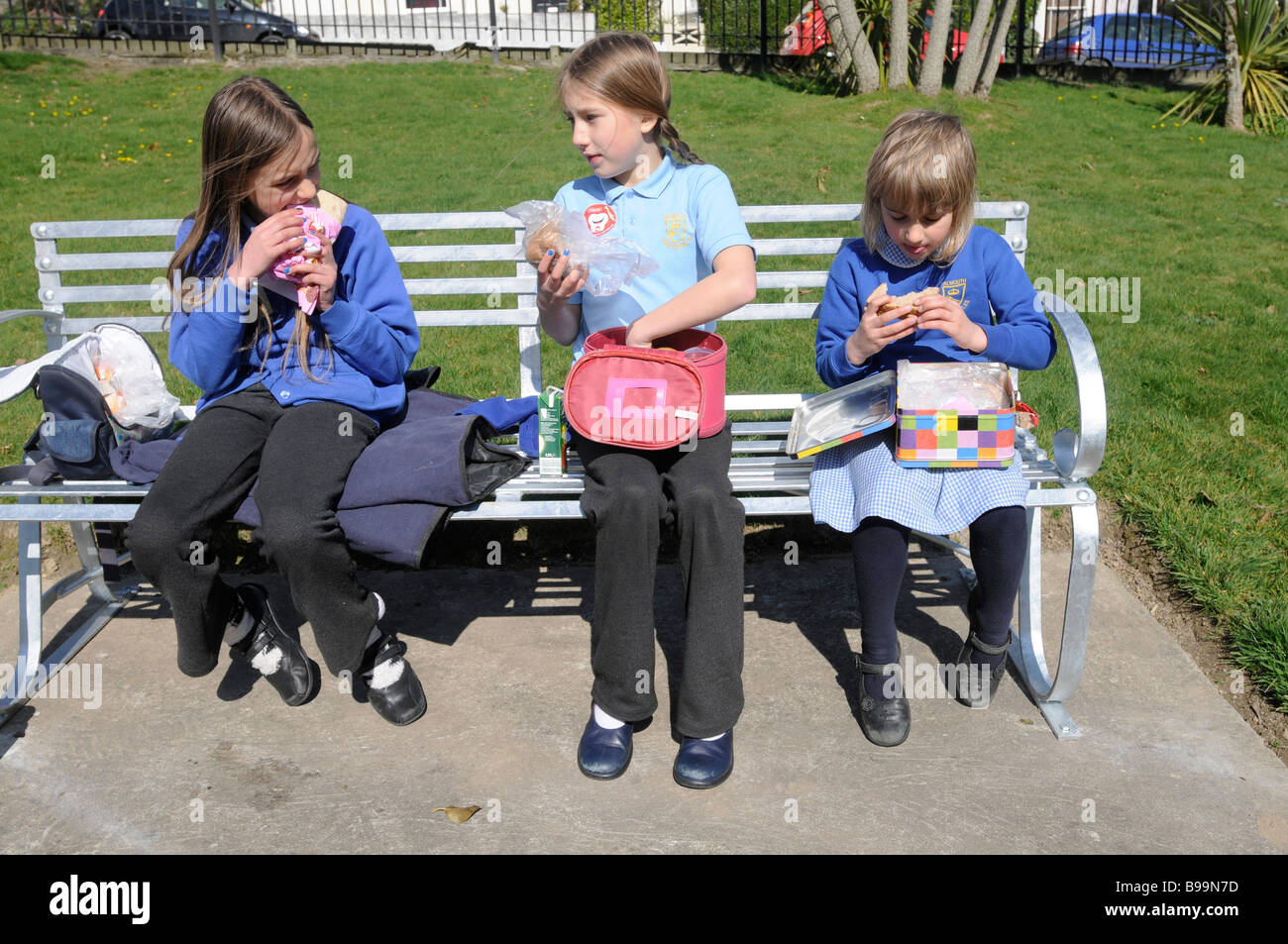 3 Schwestern in Schuluniform, die ihr Lunchpaket auf einer Parkbank Stockfoto