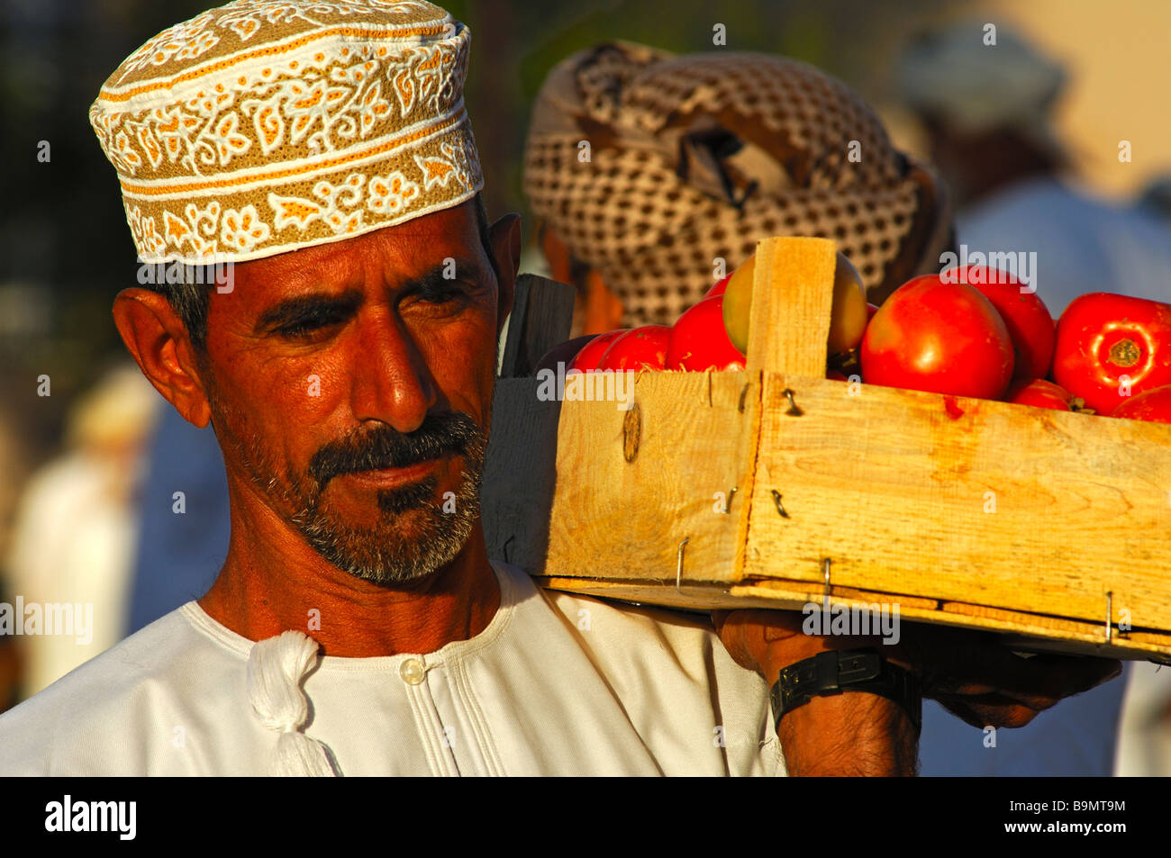 Eine omanische Gemüsehändler mit einer Kummah Kappe auf dem Kopf trägt eine Kiste voller Tomaten, Nizwa, Sultanat von Oman Stockfoto