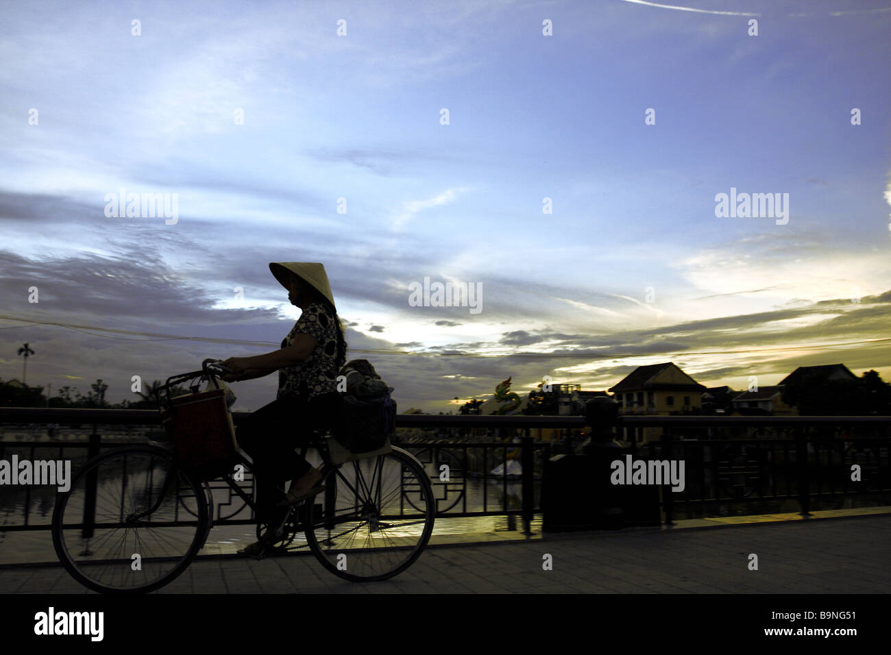 Straßenszene. Frau mit der traditionellen konische Hut Reiten ihr Fahrrad bei Sonnenuntergang auf einer Brücke in Hoi An. Vietnam Stockfoto