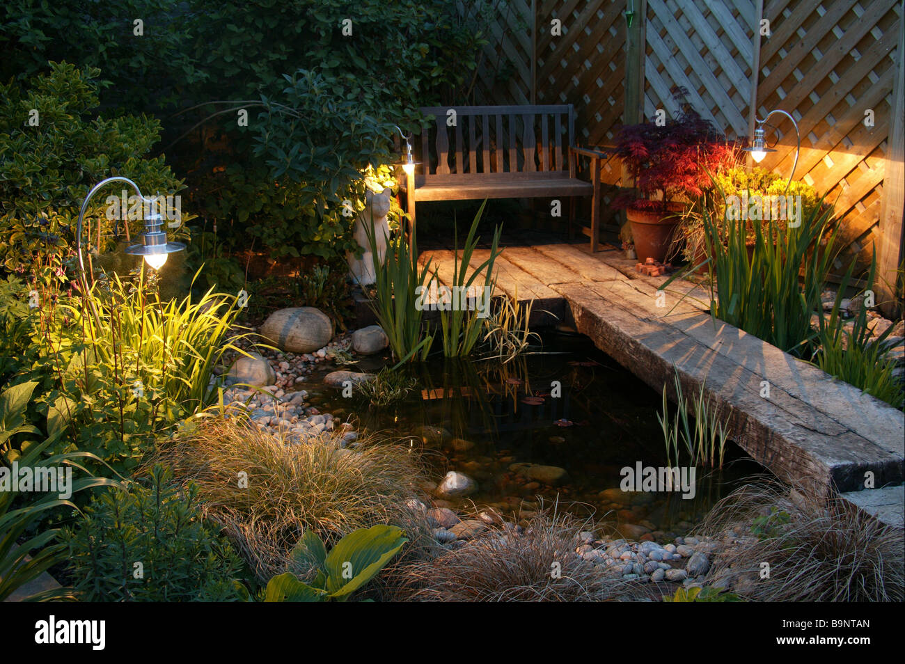 Am frühen Abend britischen Garten im Sommer, verfügt über einen Teich, Gartenstuhl, Lichter und Stein Detail. Stockfoto