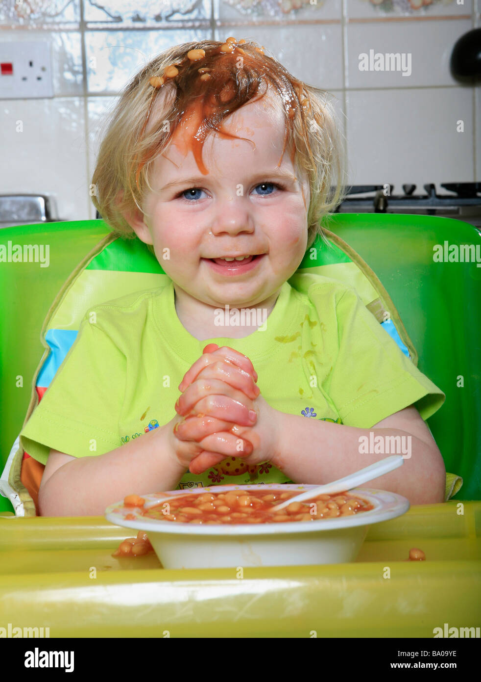 Kind mit gebackenen Bohnen auf Kopf lächelnd. Kind essen gebackene Bohnen Kind im Hochstuhl Kind in Küche Stockfoto