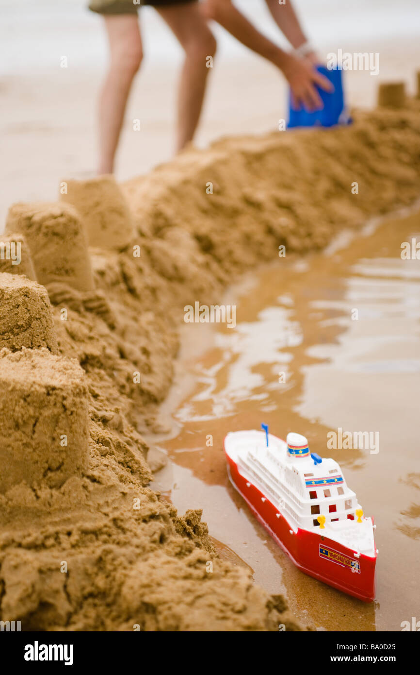 Spielzeug Schiff schwimmt in einem Graben eine Sandburg mit den Beinen und  Armen eines Mannes im Hintergrund mehr Türme bauen Stockfotografie - Alamy