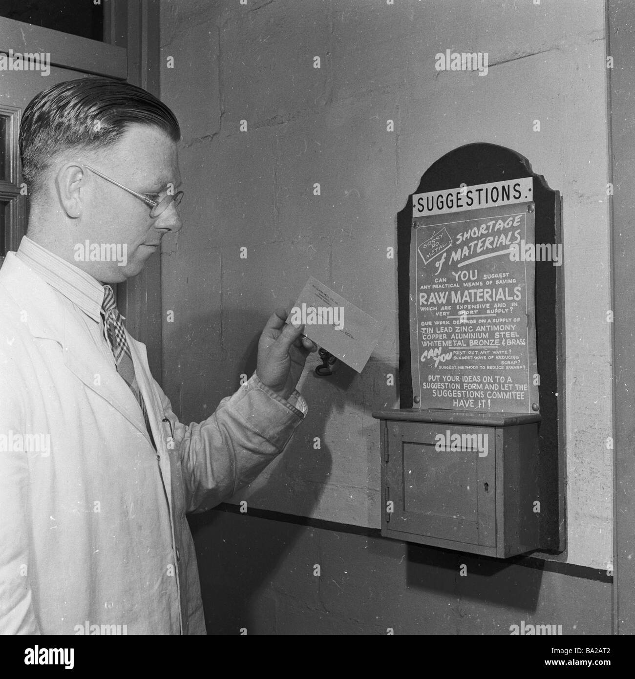 1950s, ein historischer, männlicher Angestellter, der einen weißen Mantel über einem Hemd und einer Krawatte trägt, legt eine Karte in die an der Wand montierte Vorschlagsbox der Fabrik, England, Großbritannien. Stockfoto