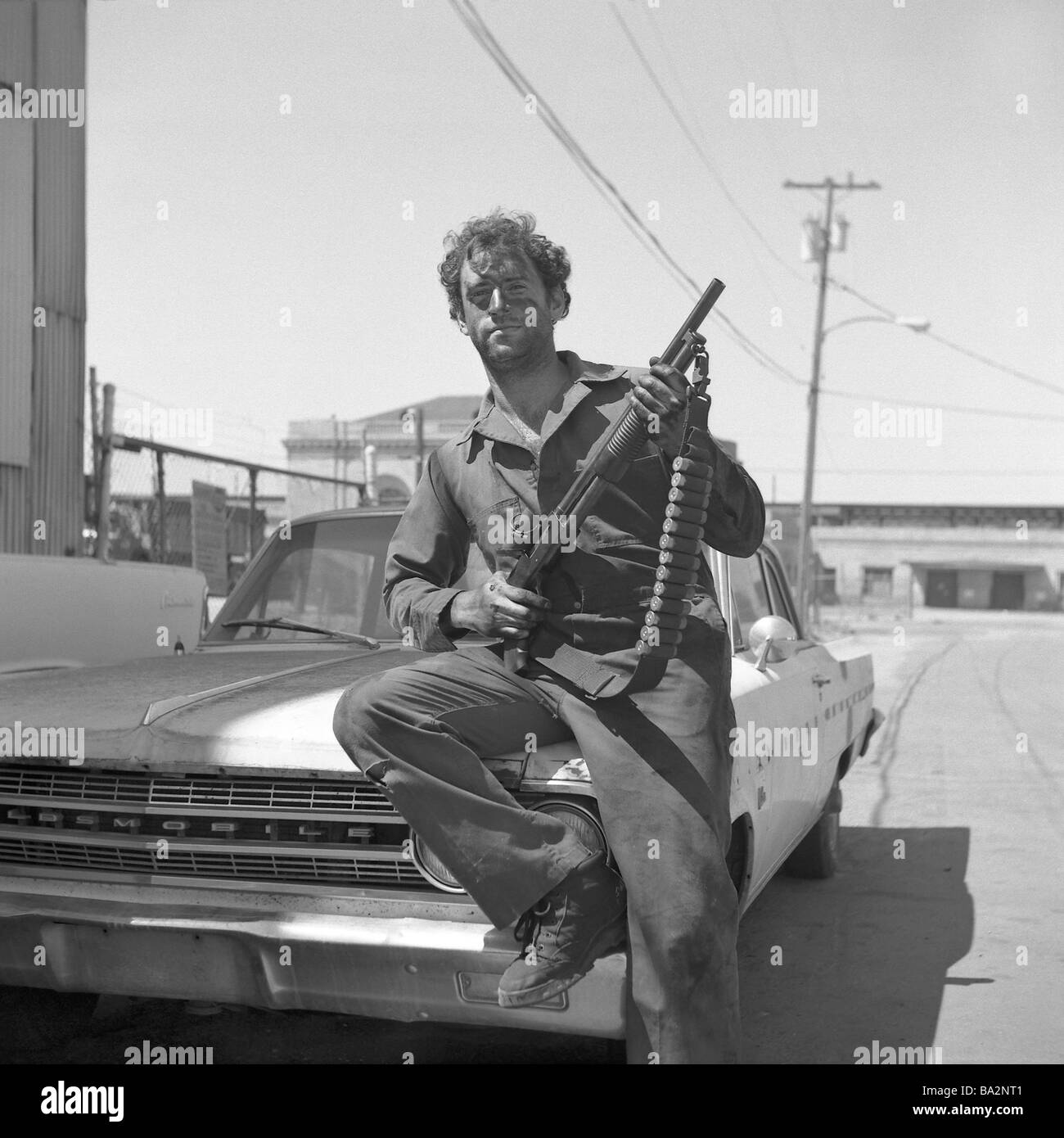 Mann Pumpgun Auto Oldtimer Motorhaube sitzt s/w Menschen Sicherheit Leibwächter Gangster schmutzigen Overall lässig cool Sonne Glas Waffe Stockfoto