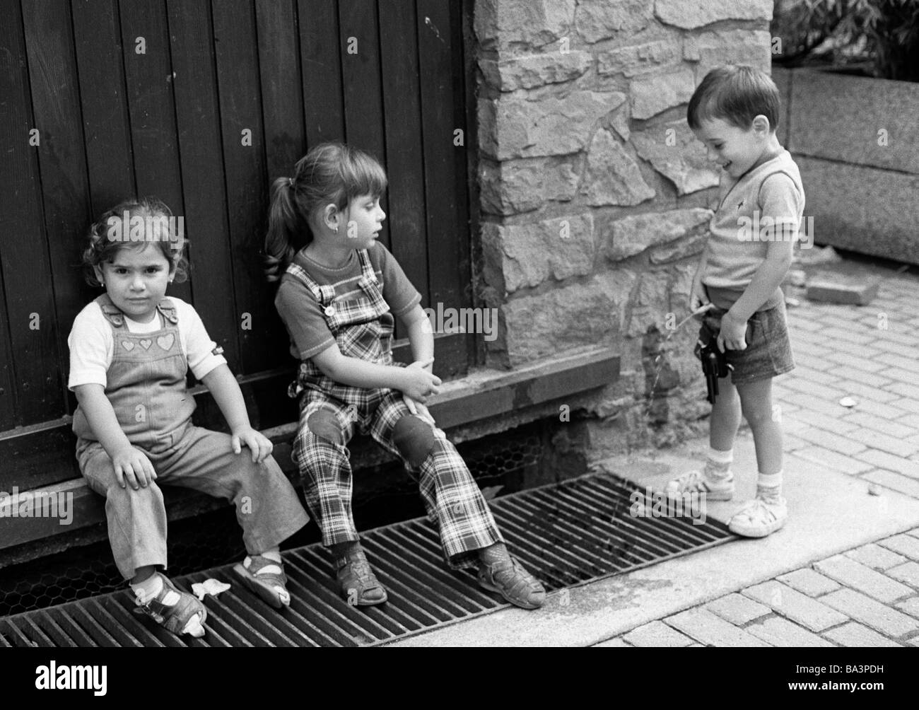 Schwarz / weiß Foto, Humor, Menschen, Kinder, zwei kleine Mädchen sitzen vor einer Haustür die Füße auf einen Abfluss der siebziger Jahre Rost, kleine Junge steht dicht neben und pinkelt in die Drain-Rost, im Alter von 4 bis 6 Jahre Stockfoto