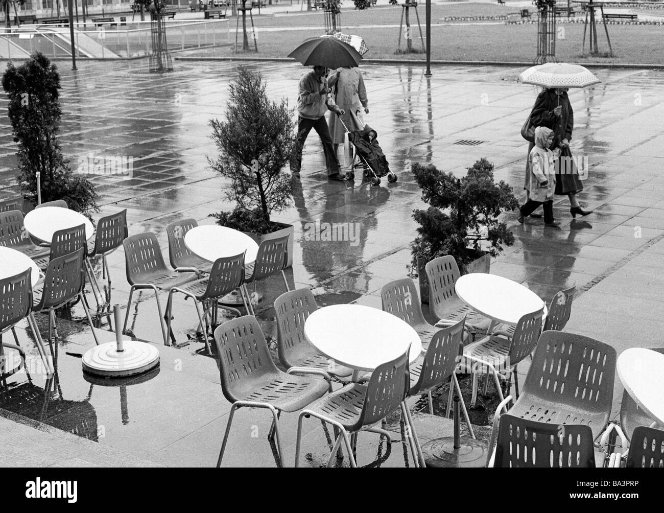 Achtziger Jahre, schwarz / weiß Foto, Menschen verpflichten sich, einen Spaziergang im Regen, Schirme, Straßencafé, leere Stühle und Gerichte Stockfoto