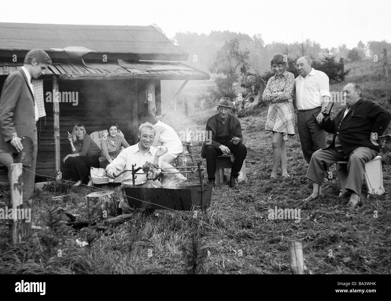 70er Jahre, schwarz / weiß Foto, Menschen, Freizeit, Grill, junge Mann dreht einen Braten auf dem Holzkohlegrill, junge und ältere Gäste schauen sehr interessiert, im Hintergrund eine Holzhütte Stockfoto