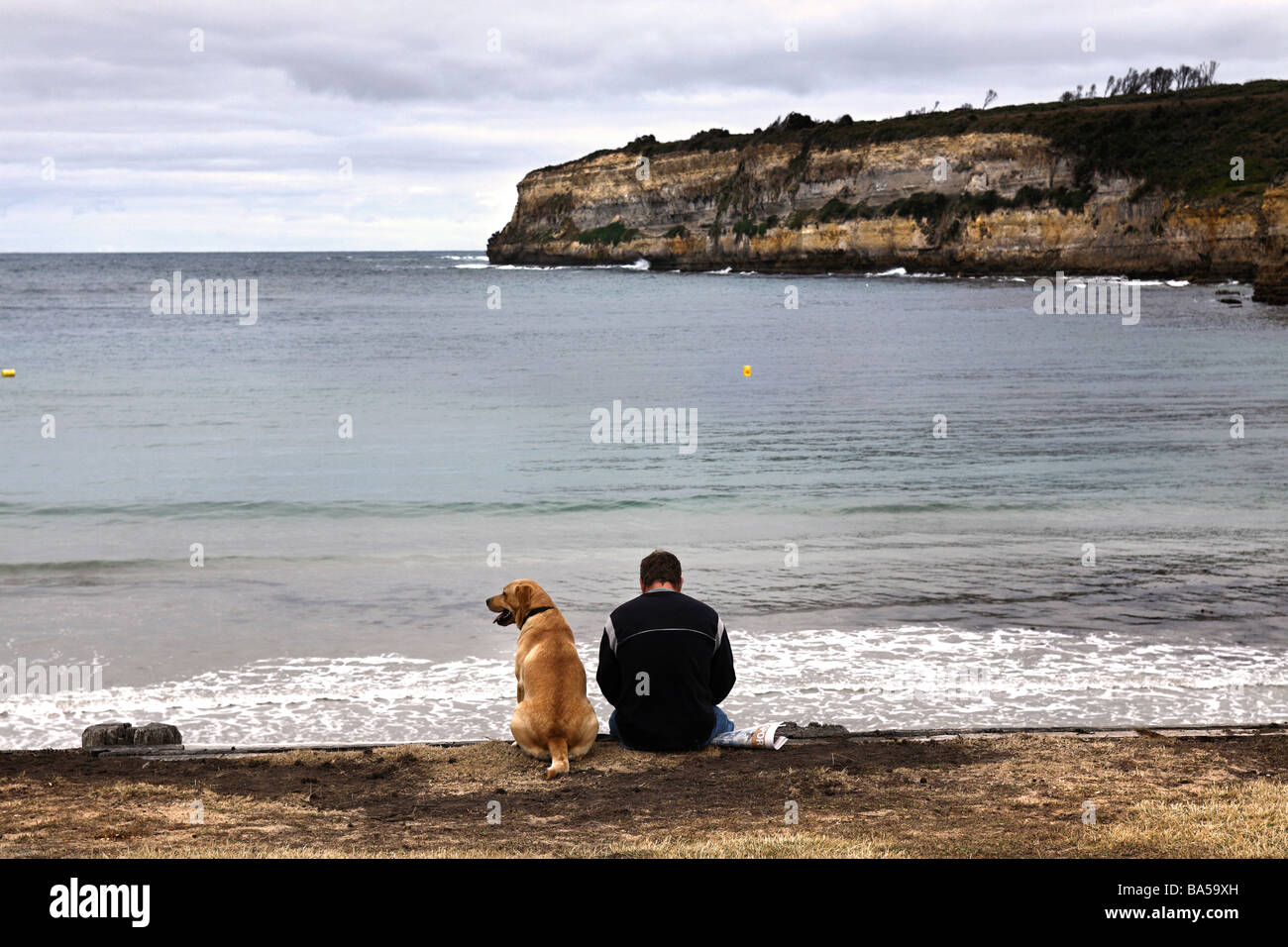 Mann und seinem Labrador Hund sitzen am Rand Wassers von Port Campbell Bay, Victoria, Australien Stockfoto