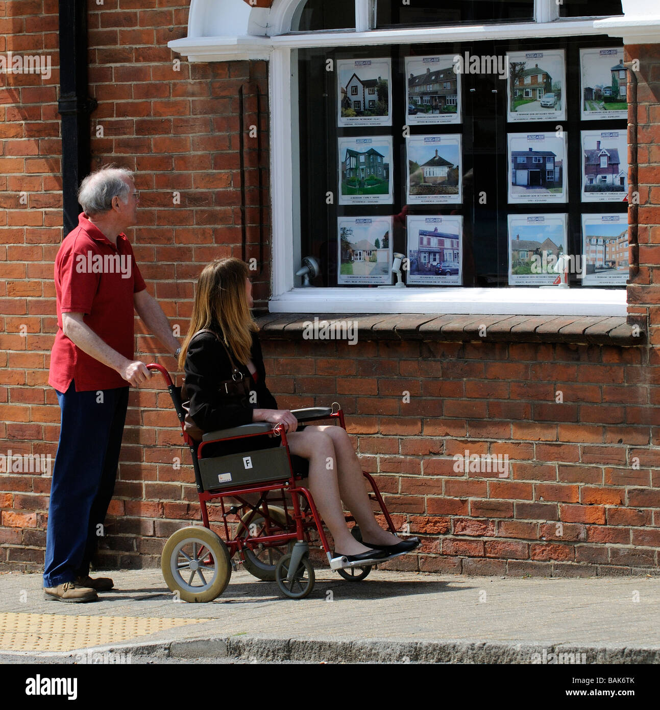 Männliche Pflegekraft schiebt Rollstuhl und weibliche Benutzer betrachten Immobilien zum Verkauf in Immobilienmakler Fenster Stockfoto