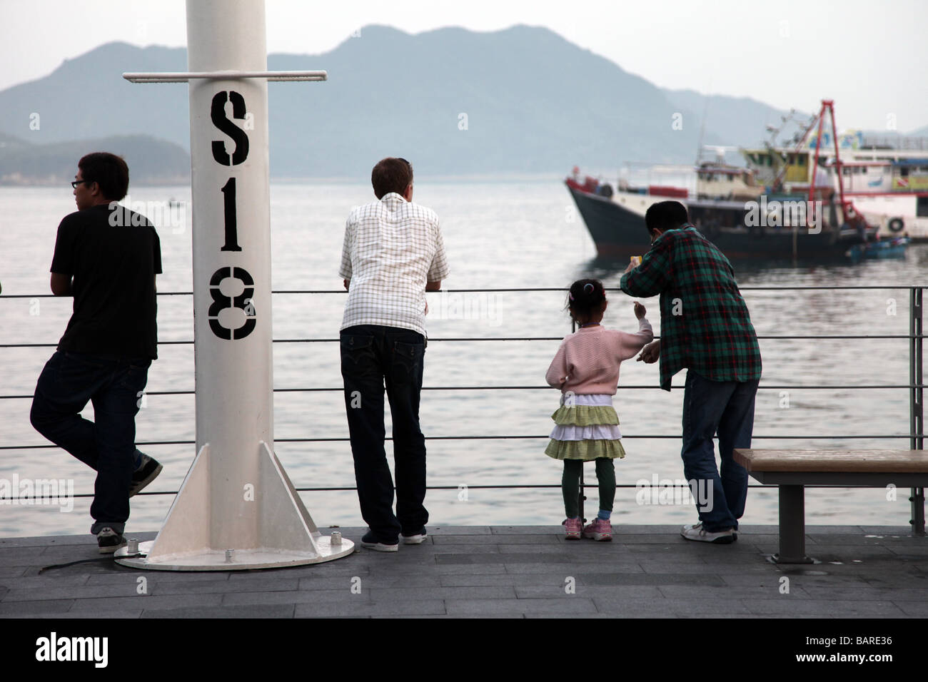 4 Personen mit Blick auf das Meer aus dem Dock eines Hafens. Es ist ein Boot auf das Meer und die Berge im Hintergrund bei Tageslicht Stockfoto