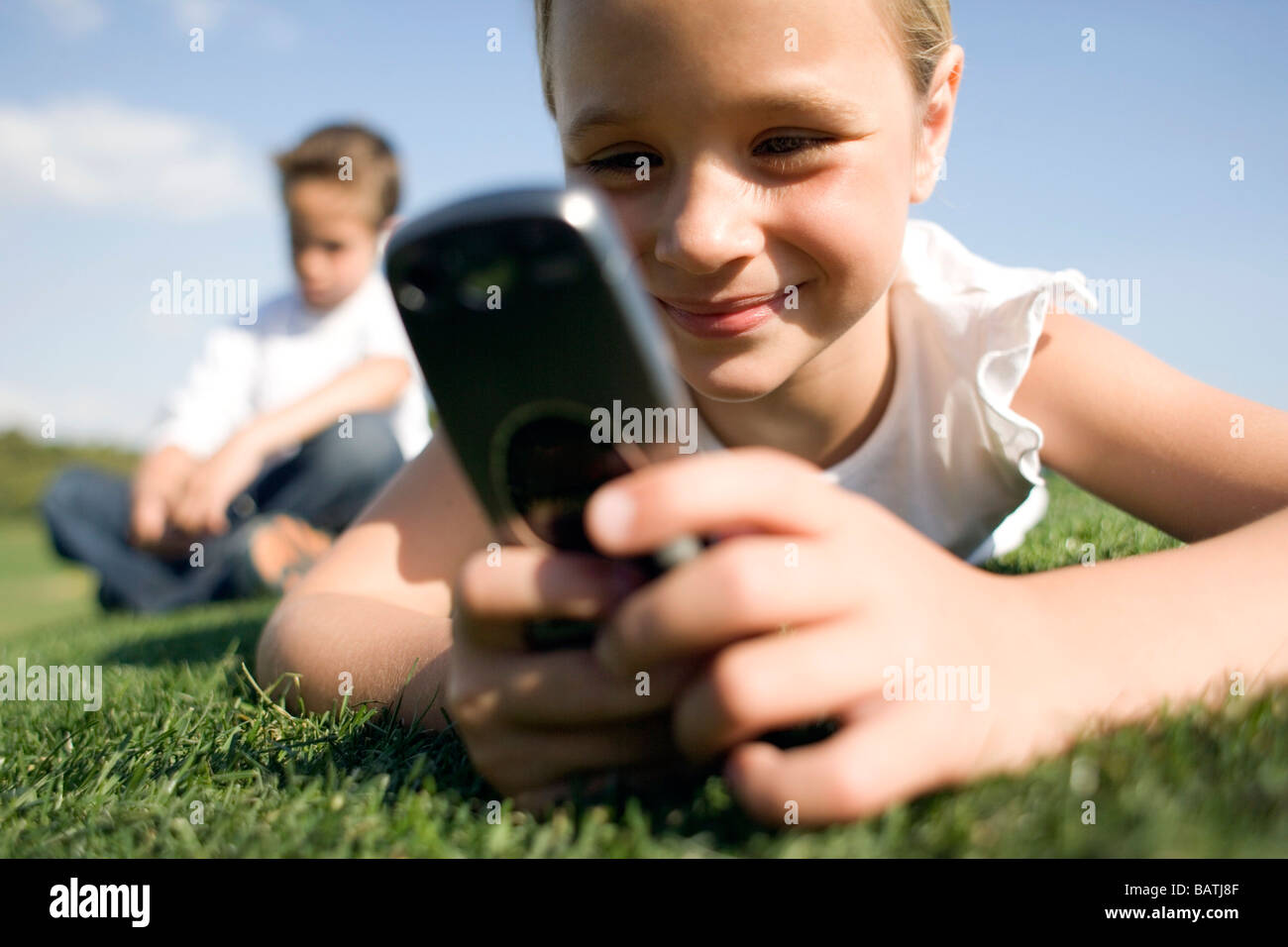 Mädchen mit einem mobilen Phonewhile auf dem Rasen liegen. Ein Junge sitzt in Thebackground. Stockfoto