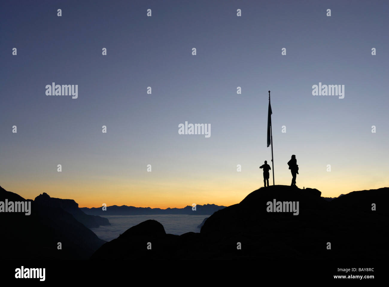 zwei Wanderer auf Kuppel mit Fahnenmast, Morgendämmerung über Nebelbank im Tal Gschnitztal, Kirchdachspitze im Hintergrund, Bremer Huette, Stuba Stockfoto
