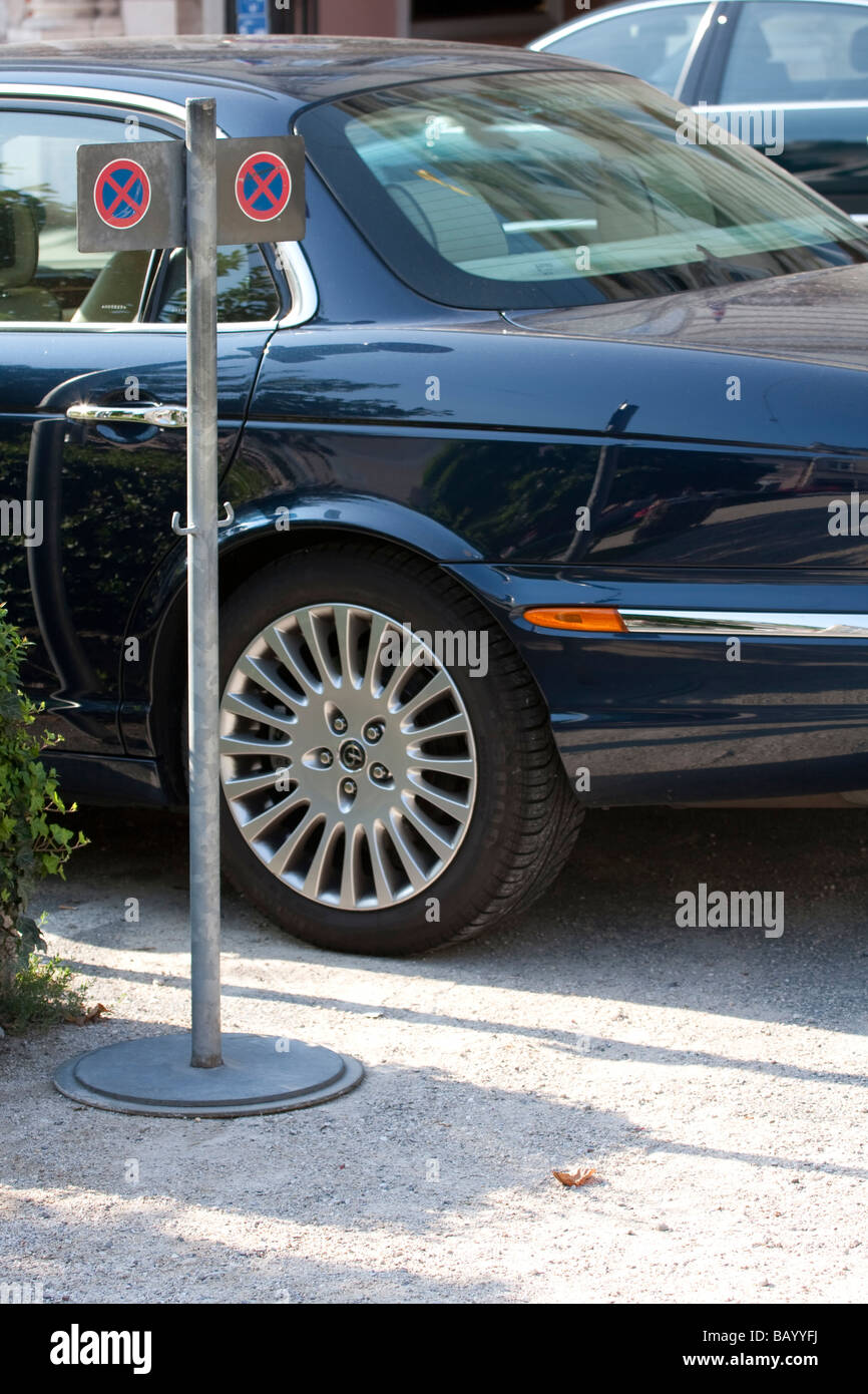 Ein metallisches Parkverbot-Schild in der Nähe eines geparkten dunkelblauen Autos mit poliertem Rad, das sich auf einer Kiesoberfläche unter Sonnenlicht befindet. Stockfoto