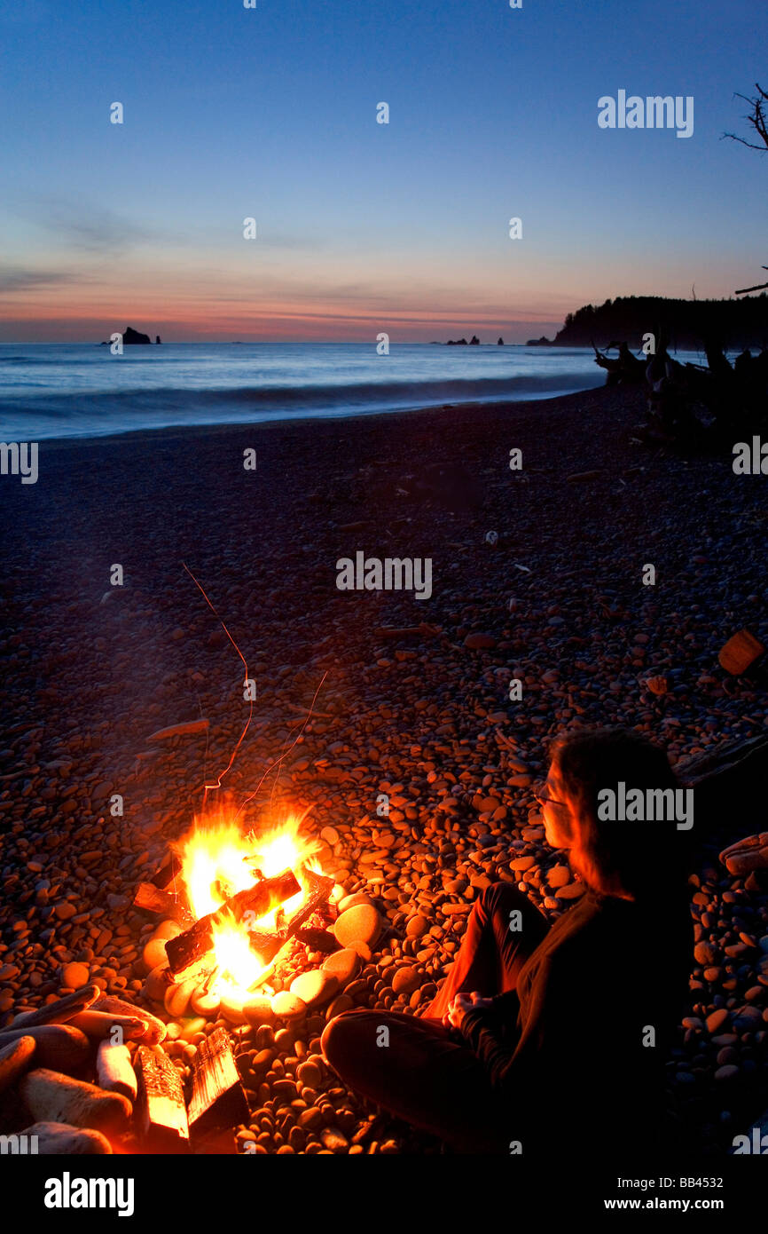 USA, Washington, Olympic Nationalpark, Rialto Beach. Frau genießt Lagerfeuer am Strand bei Sonnenuntergang. Stockfoto