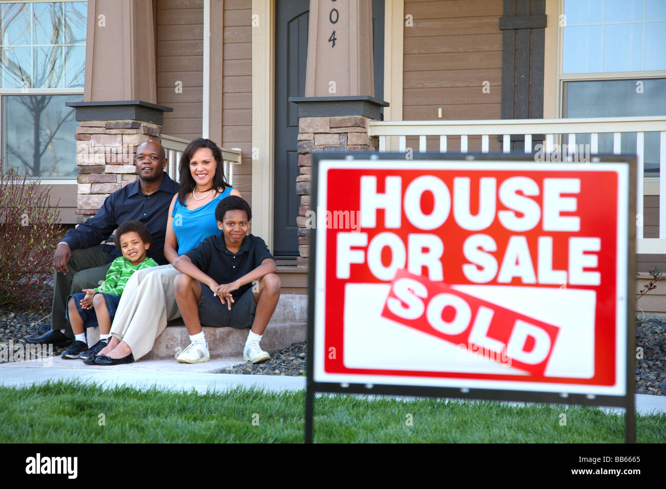 Afroamerikanische Familie sitzen zusammen auf der Eingangstreppe des Hauses mit Immobilien Schild im Vordergrund Stockfoto