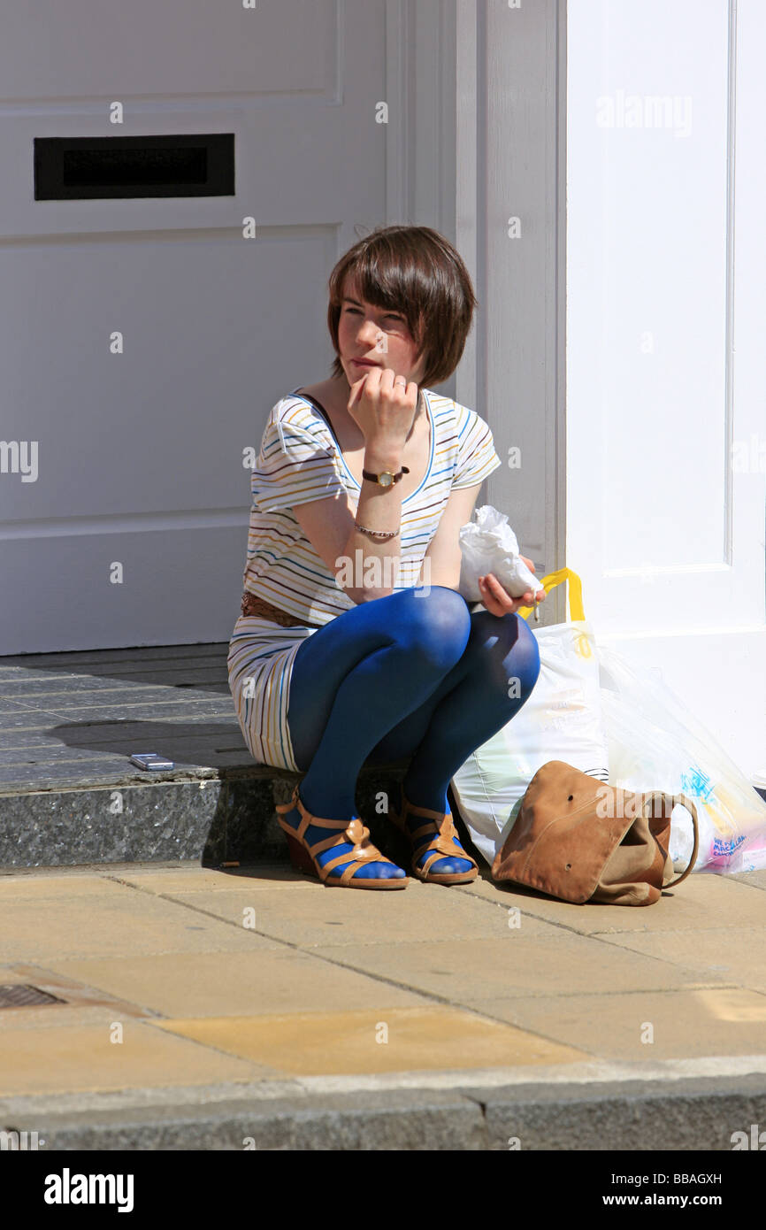 Weibliche Teenager sitzen auf einer Haustür essen Süßigkeiten aus der Tasche, während sie darauf warteten, mit ihren Einkaufstaschen für eine Kabine Stockfoto
