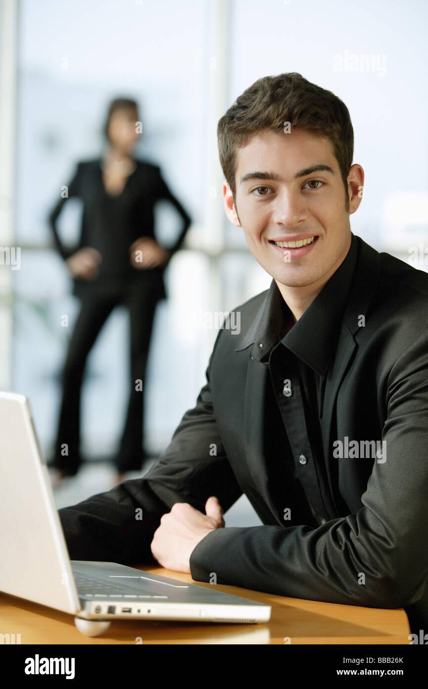 Männliche Executive in schwarz gekleidet, sitzt neben Laptop, Blick in die Kamera, Lächeln Stockfoto
