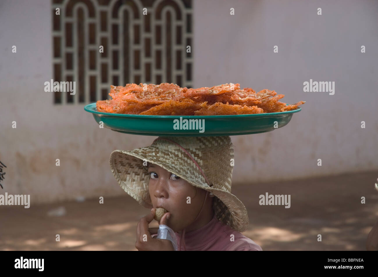 Kambodschanischen Straßenverkäufer balanciert eine Pfanne mit frittiertem Brot und Garnelenkuchen (ប្រហិតបង្គាបំពង), während er eine Litschi isst. Stockfoto