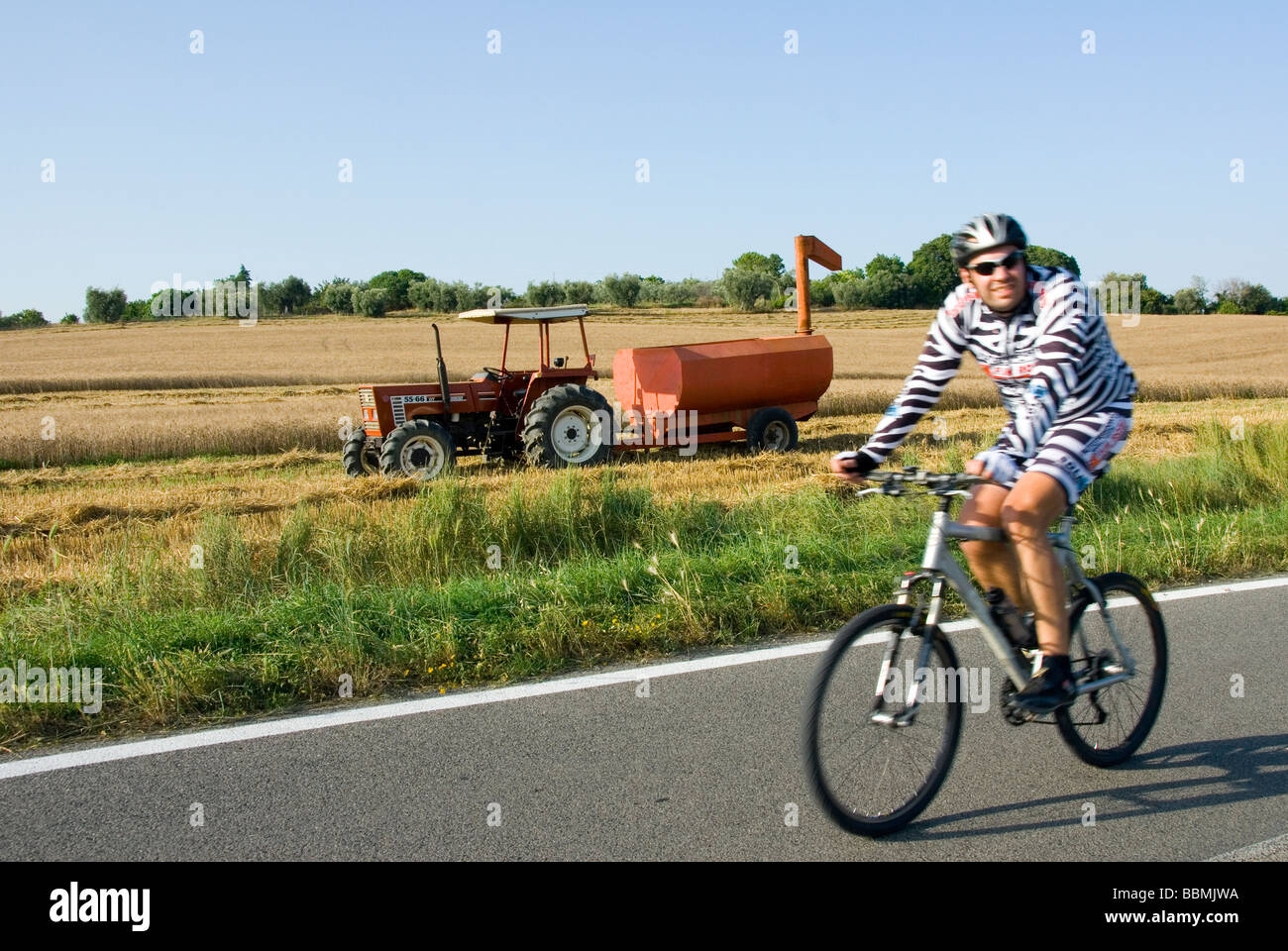 Radfahrer in Zebra Radsport Outfit reitet auf der Straße hinter einem Traktor und Mähdrescher in der Nähe von Magliano in Toscana, Maremma Italien Stockfoto