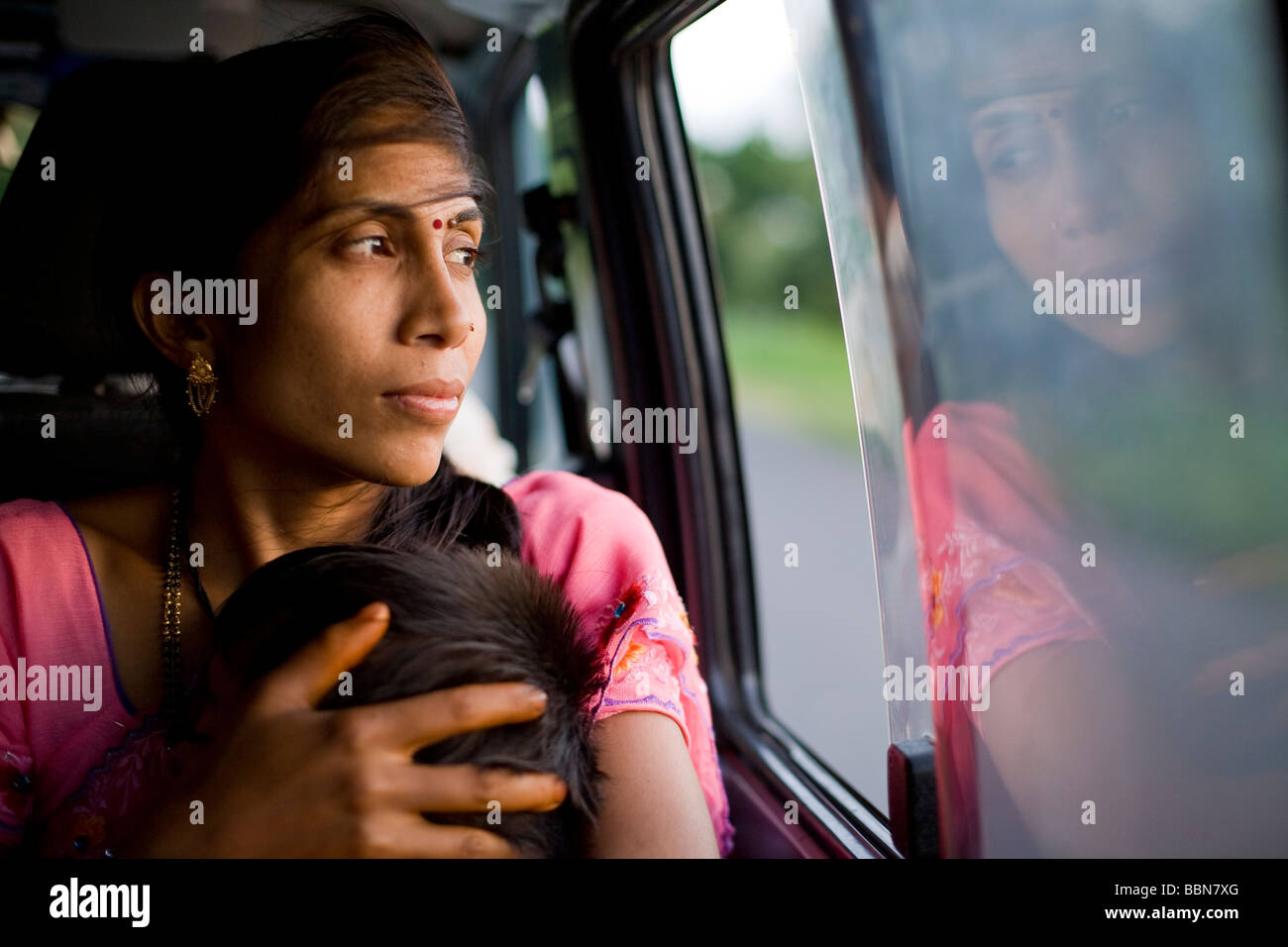 Indische Mutter spiegelt sich in dem Autofenster wie sie ihren schlafenden Sohn auf der langen Autofahrt aus ihrem Dorf in die Stadt wiegen. Stockfoto
