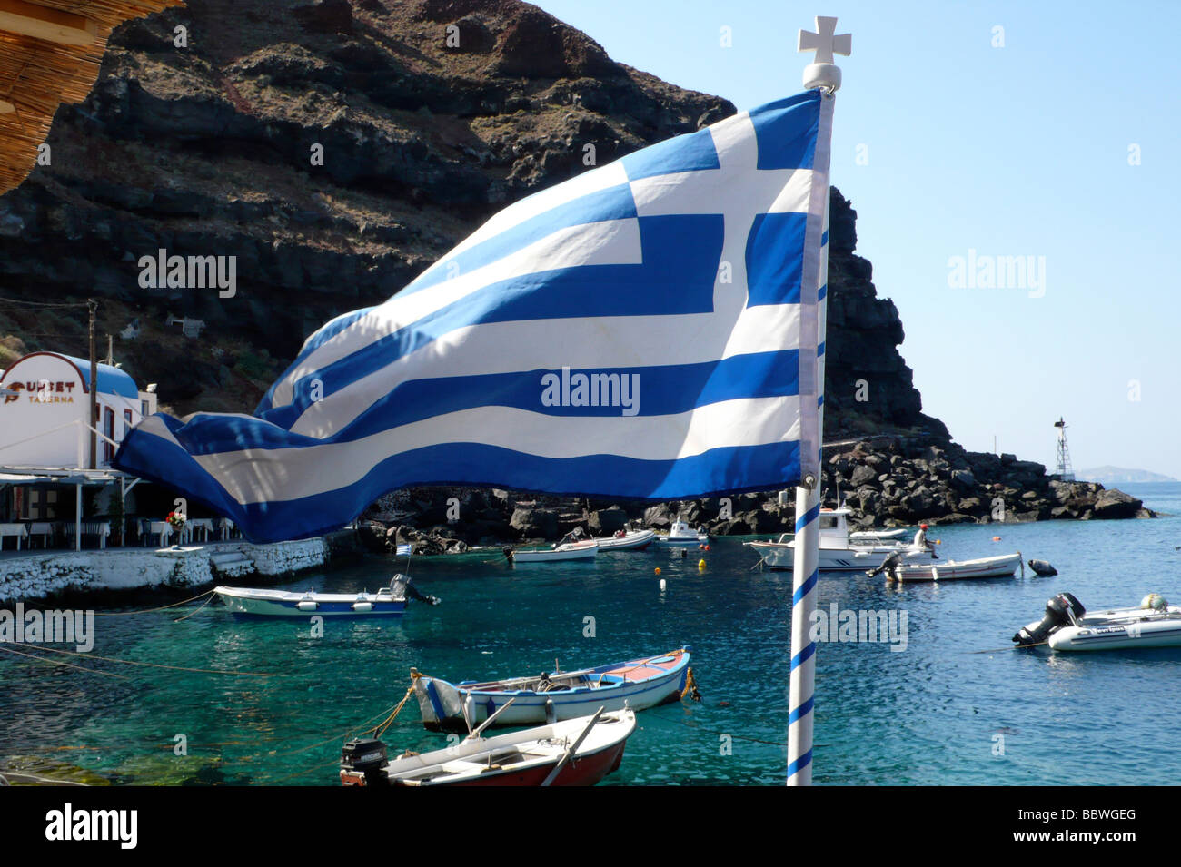 Griechische Flagge am Ammoudi - in der Nähe von Oia mit Fischerbooten im Hintergrund Stockfoto