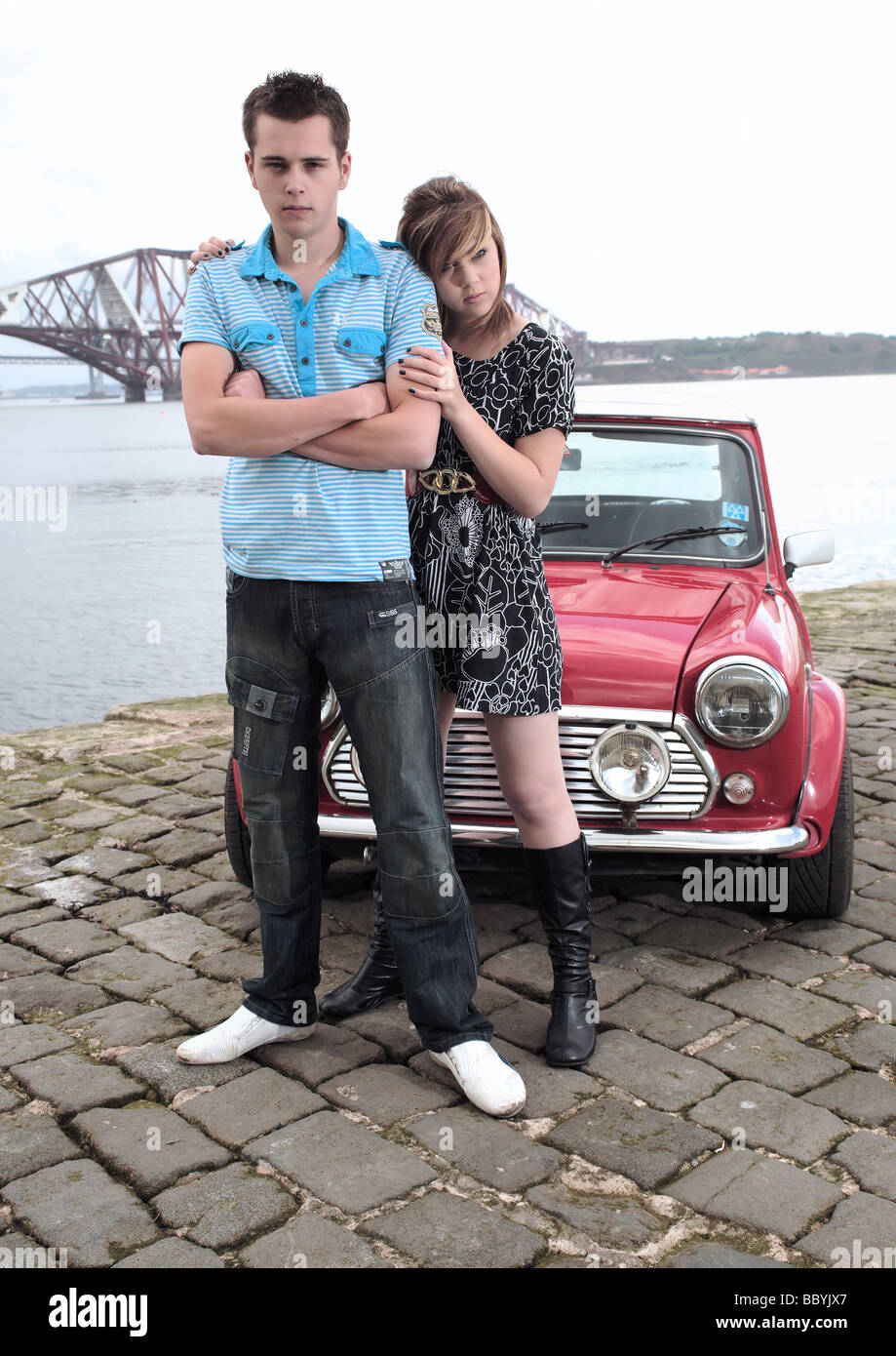 Junge Junge und Mädchen Models im Bild mit einem roten Mini Cooper auf einem Pier, überragt von dem her Schiene Brücke in der Nähe von Edinburgh. Stockfoto