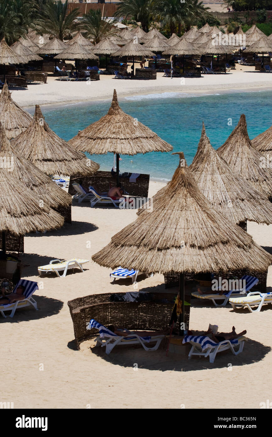 Urlauber sitzen unter reetgedeckten Sonnenschirmen auf einer exotischen Bäumen gesäumten Palmenstrand Stockfoto