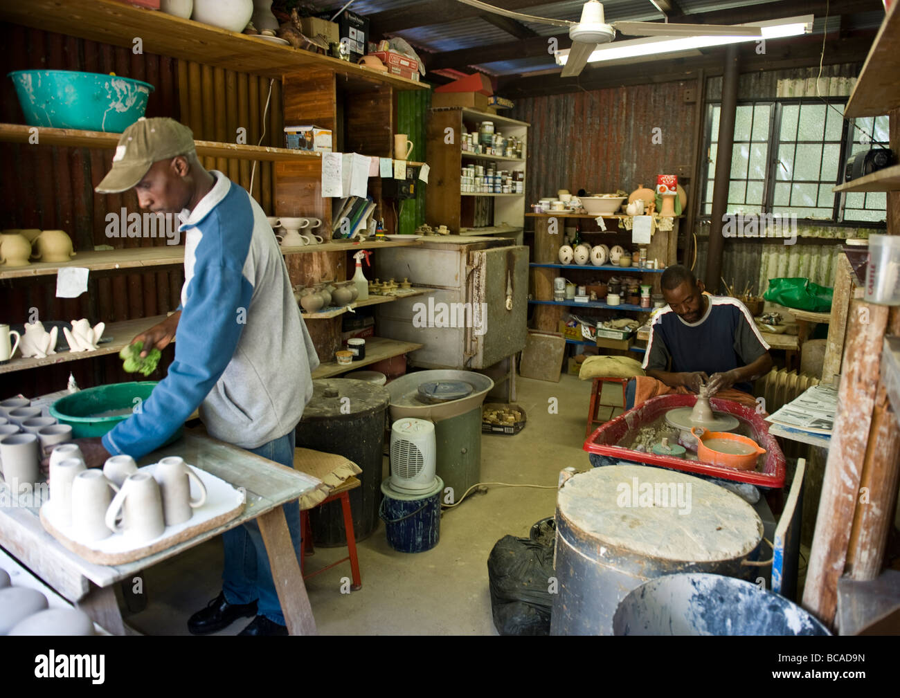 Afrikanische Künstler gestalten eine Tasse auf der Töpferscheibe in seiner Werkstatt und ein anderer Mann, Vermittlung von Keramik zum Verkauf im Souvenirladen. Stockfoto