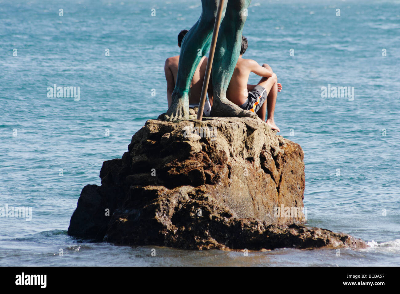 Zwei spanische Jungs sitzen auf Felsen am Fuße des Neptun-Statue im Meer vor der Küste von Gran Canaria Stockfoto