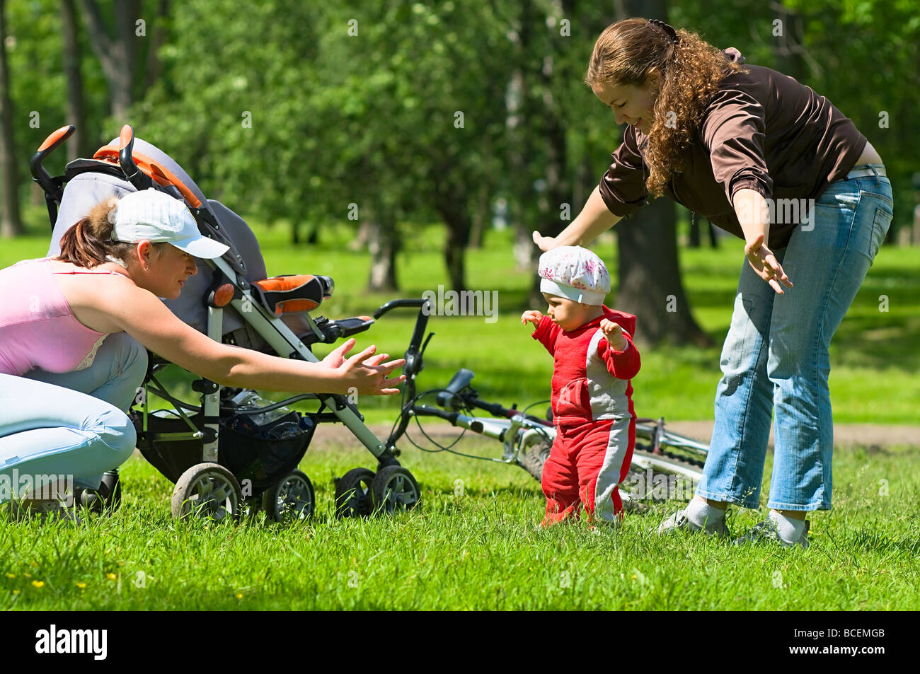 Zwei Frauen helfen, Kleinkind, erste Schritte im Park zu tun. Grünen Rasen und Laub rund um sie. Es ist ein Sommer. Kinderwagen und Fahrrad Stockfoto