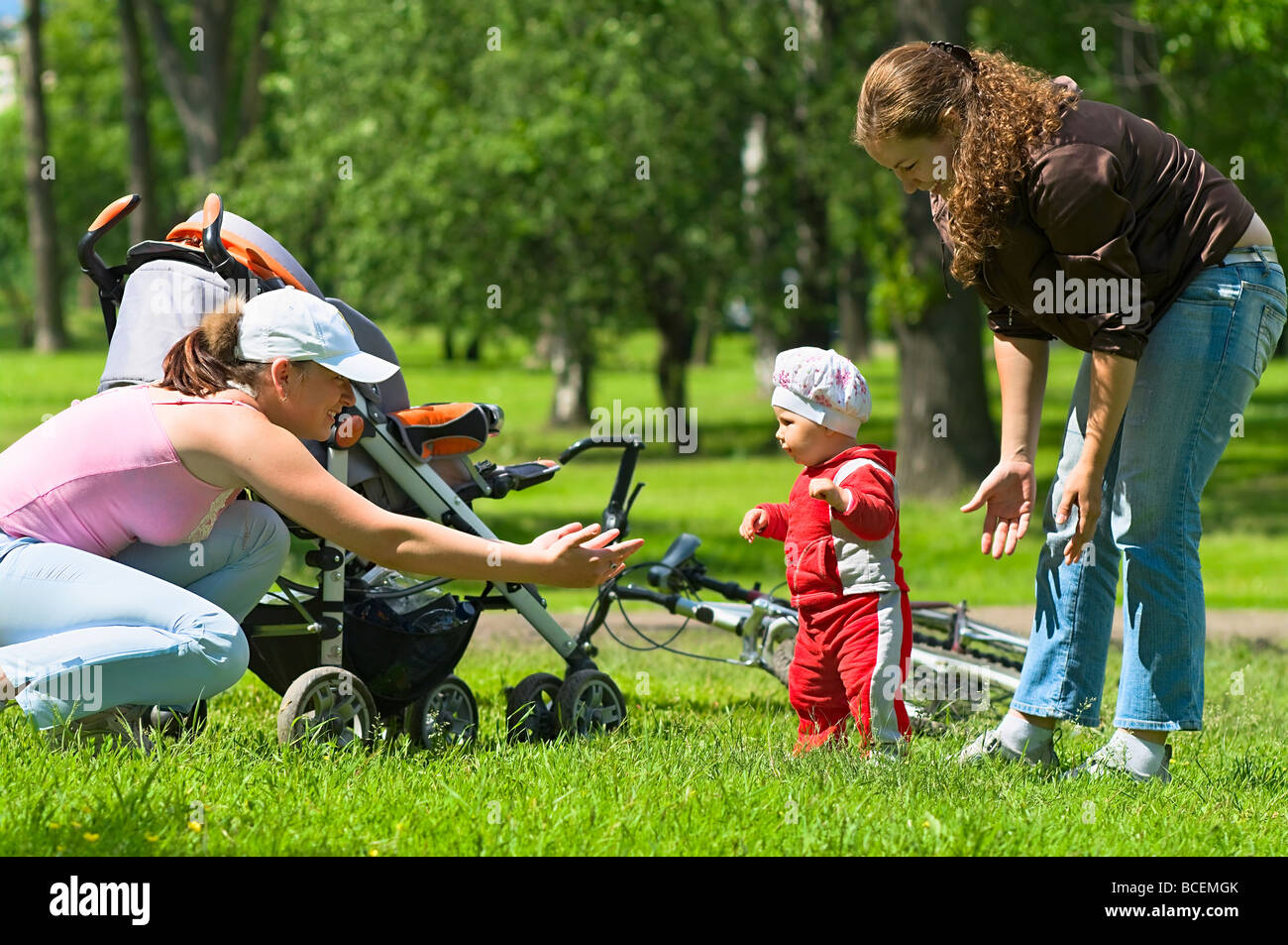 Zwei Frauen helfen, Kleinkind, erste Schritte im Park zu tun. Grünen Rasen und Laub rund um sie. Es ist ein Sommer. Kinderwagen und Fahrrad Stockfoto