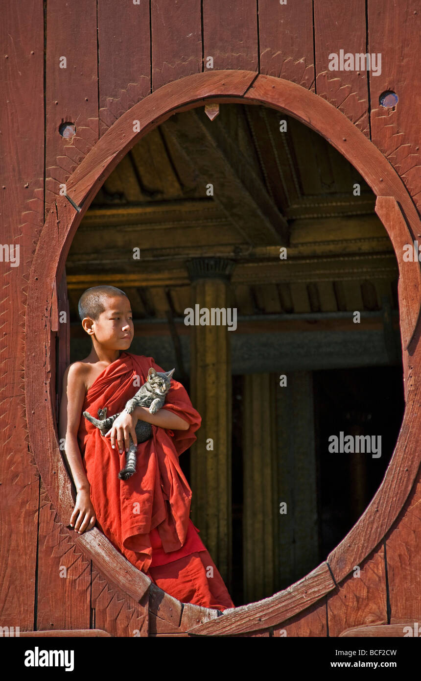 Myanmar. Burma. Nyaung Shwe. Ein junger Novize hält eine Katze am ovalen Fenster des hölzernen Shwe Yaunghwe Klosters. Stockfoto