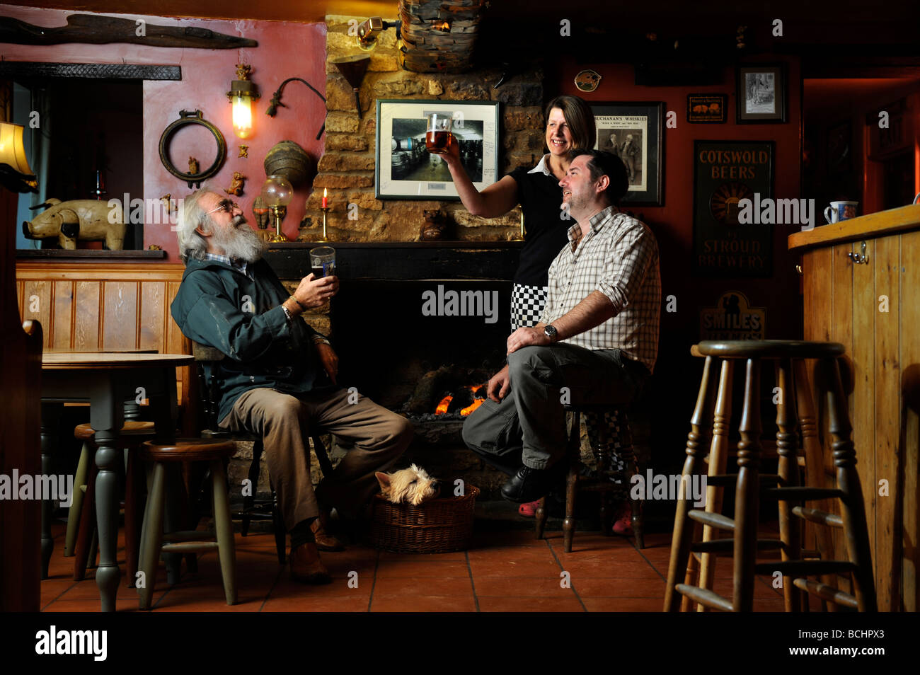 ZWEI MÄNNER IM CHAT DURCH EINE OFFENE FEUER MIT EINEM WEIBLICHEN CHEF IN EINEM TRADITIONELLEN PUB GLOUCESTERSHIRE ENGLAND UK Stockfoto