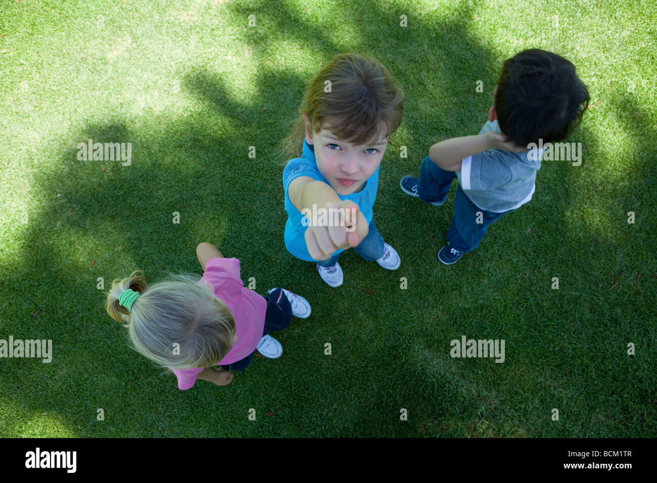 Drei Kinder stehen auf dem Rasen im Schatten, ein Mädchen in die Kamera