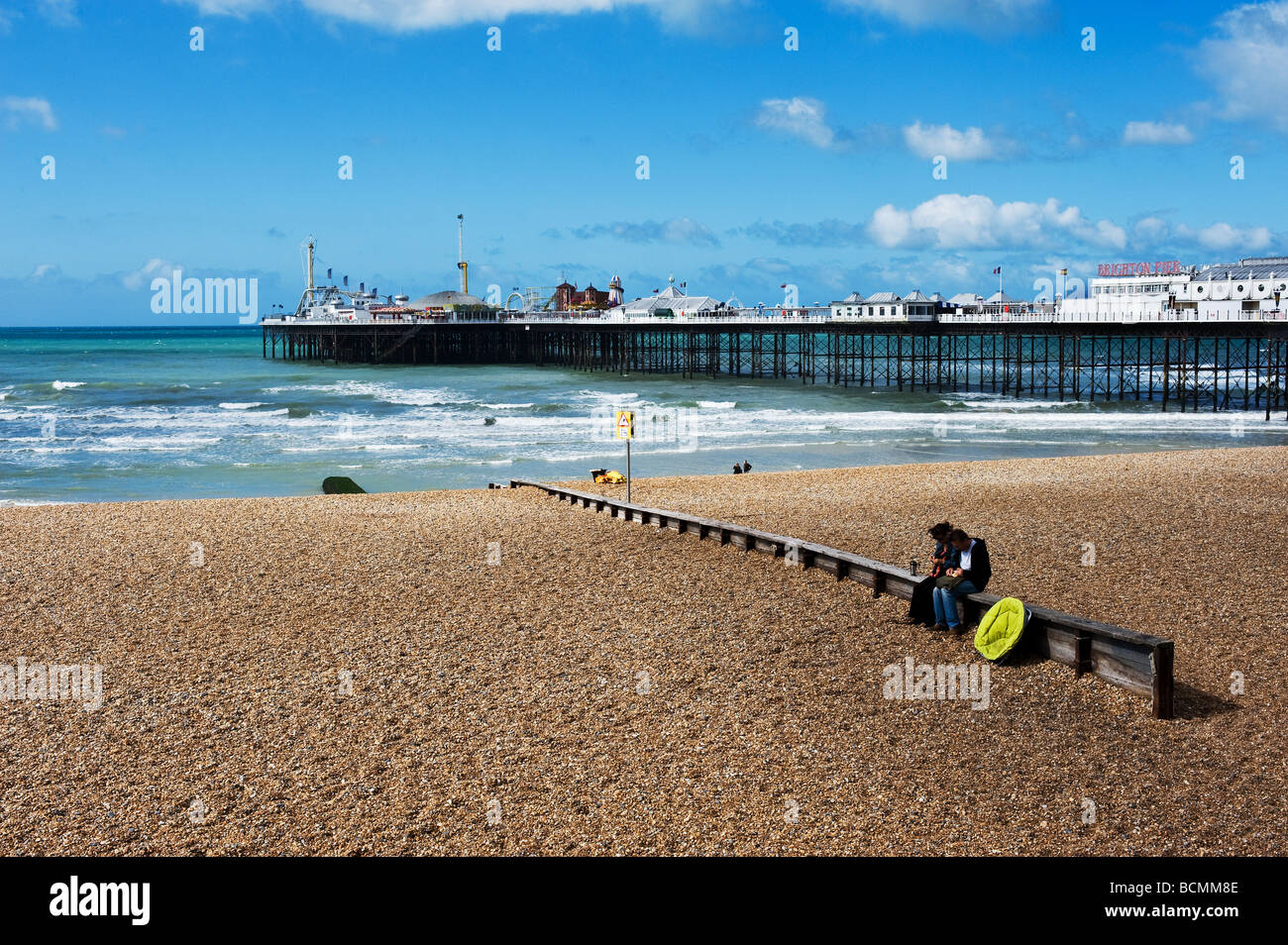 Zwei Menschen sitzen auf einem Wellenbrecher auf der Kiesstrand in der Nähe der Pier in Brighton in Sussex. Stockfoto