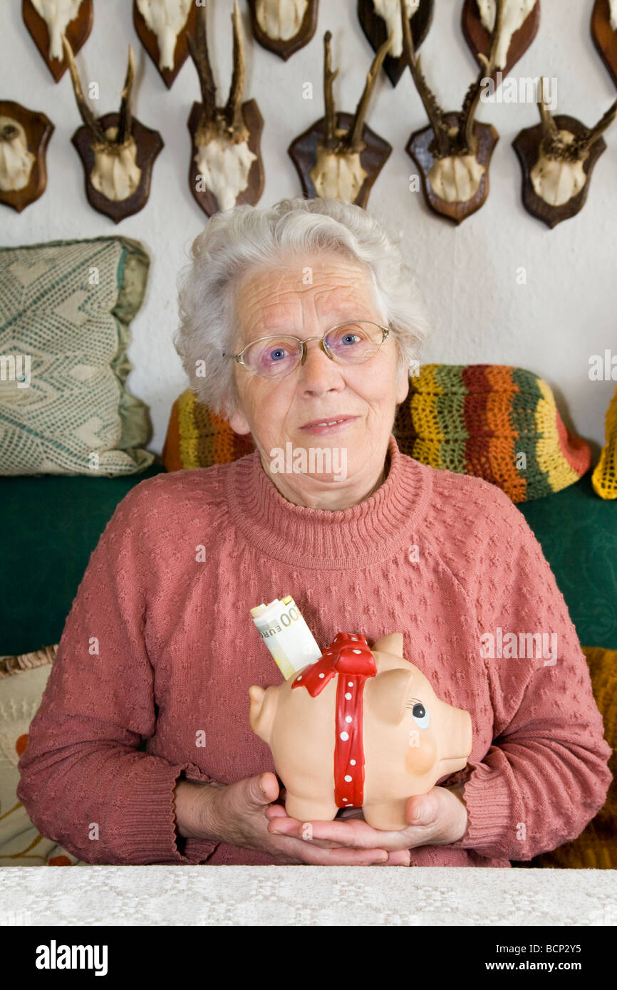 Frau in Ihren Siebzigern Sitzt Auf Einem Sofa Im Wohnzimmer Und Hält Ein Sparschwein Mit Geldscheinen Im Schlitz Stockfoto