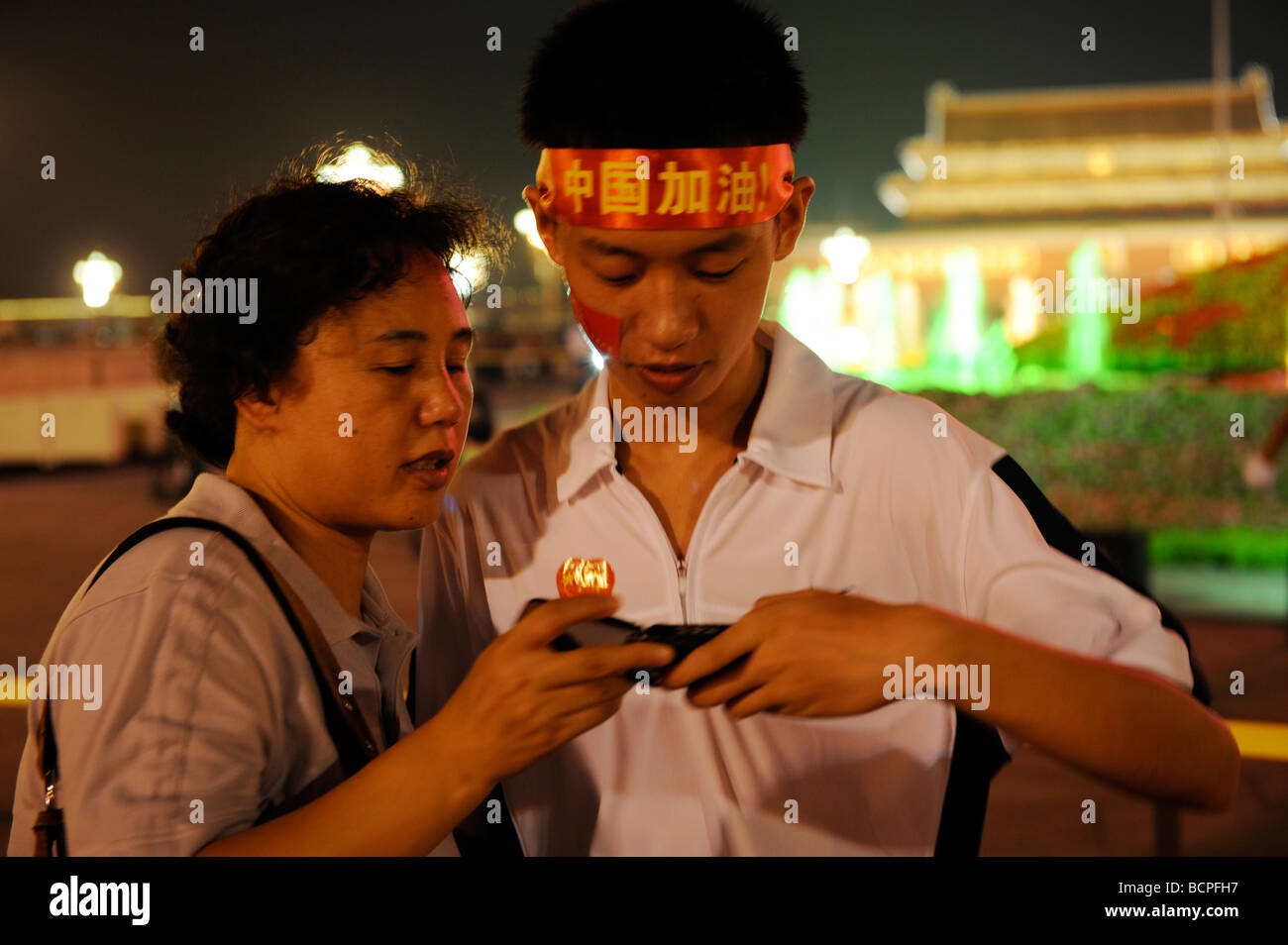 Chinesischen Mutter und Sohn im Teenageralter mit "Go China!" Stirnband Blick auf Handy in Tian An Men Square bei Nacht, Peking, China Stockfoto