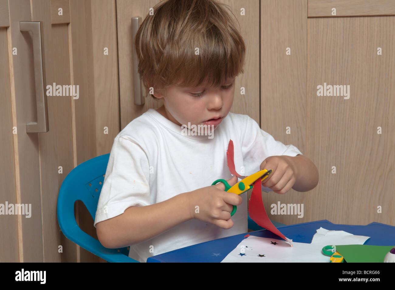 Der dreijährige Junge schneidet mit einer Schere und sitzt an einem kleinen Tisch in der Küche Stockfoto