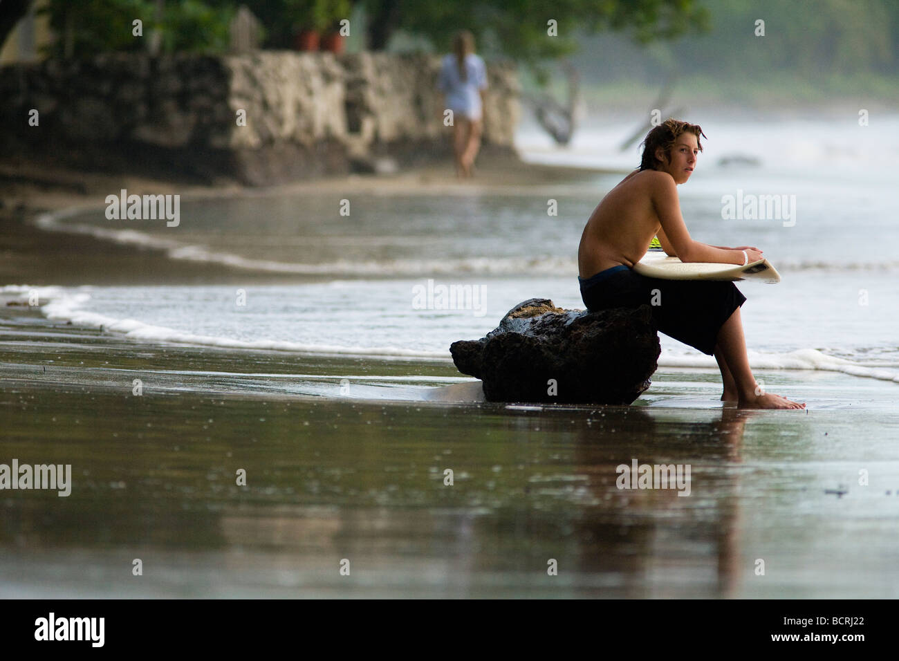 Ein junger Surfer sitzt auf einem Baumstamm auf Playa Tamarindo in Guanacaste, Costa Rica, hält sein Board und ruhen, während die Wellen warten. Stockfoto