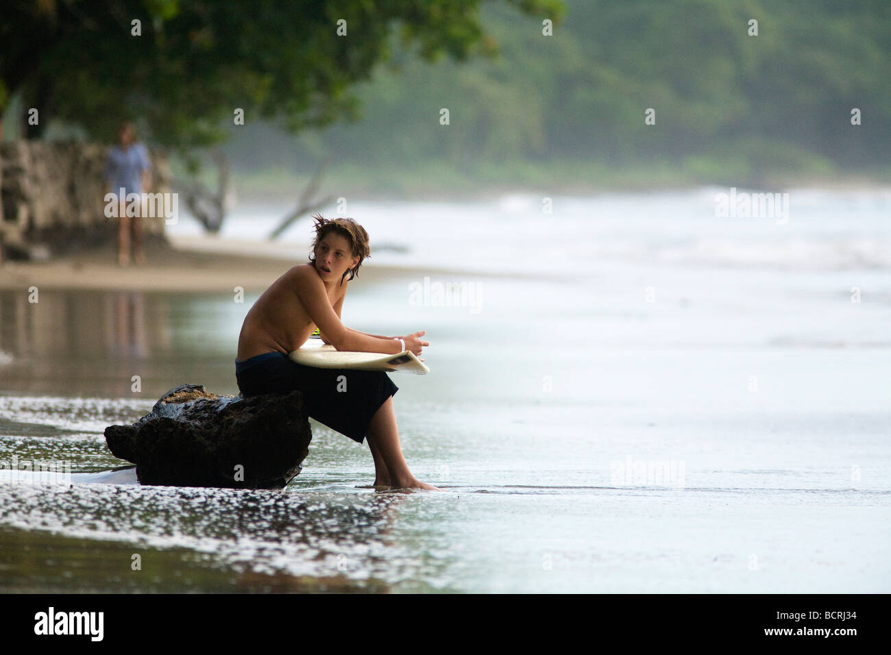 Ein junger Surfer sitzt auf einem Baumstamm auf Playa Tamarindo in Guanacaste, Costa Rica, hält sein Board und ruhen, während die Wellen warten. Stockfoto