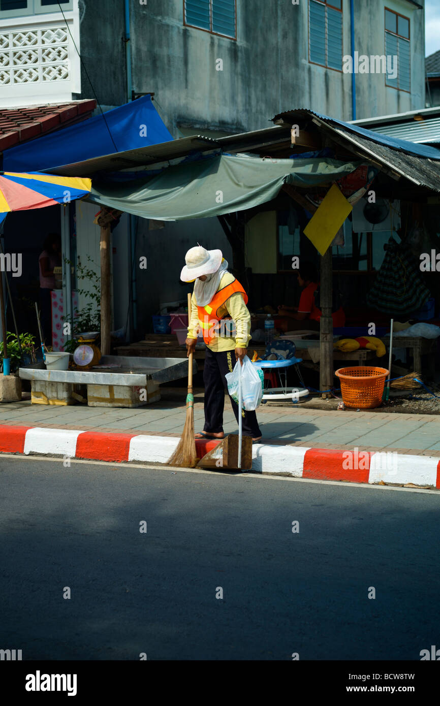 Ein Thai Street cleaner fegt eine Straße entlang einer roten und weißen Bordstein in Nathon, Koh Samui, Thailand. Stockfoto
