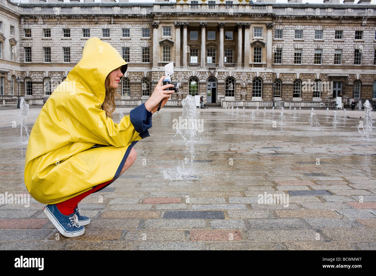 Eine junge Frau in gelb wasserdicht Mac rote Socken zu beschichten und blau Schuhe Fotografien der Brunnen im Somerset House in London Stockfoto