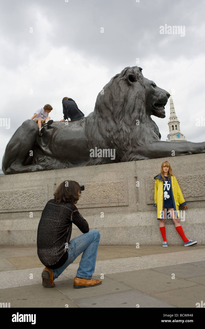 Eine junger Mann Photographsa hübsche blonde Mädchen in einem gelben Mantel und roten Socken Kniestrümpfe von den Löwen auf dem Trafalgar Square posiert Stockfoto