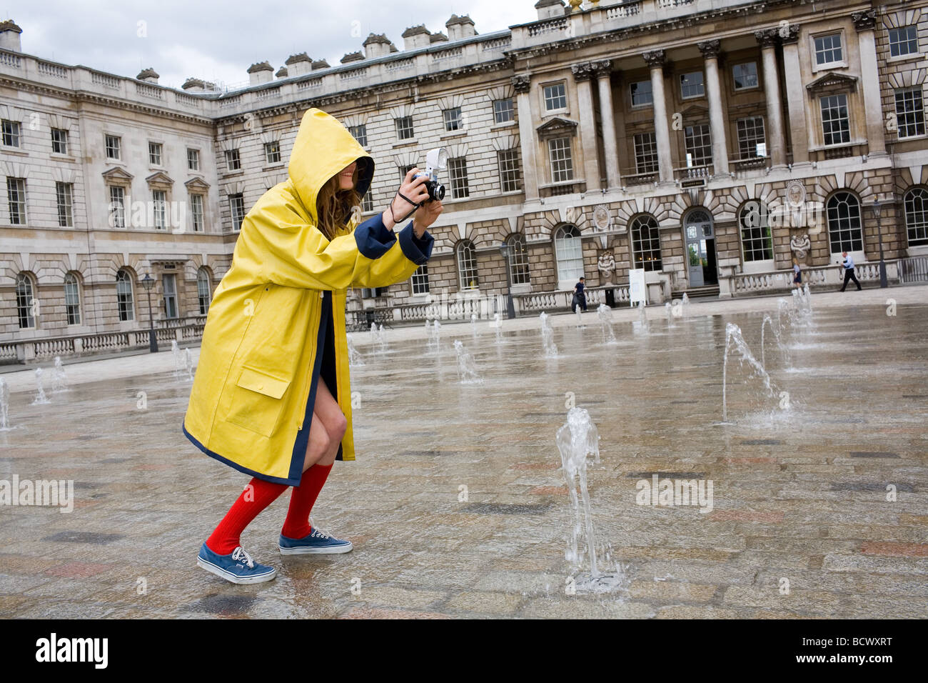 Eine junge Frau in gelb wasserdicht Mac rote Socken und blau Schuhe Fotografien der Brunnen im Somerset House in London Stockfoto