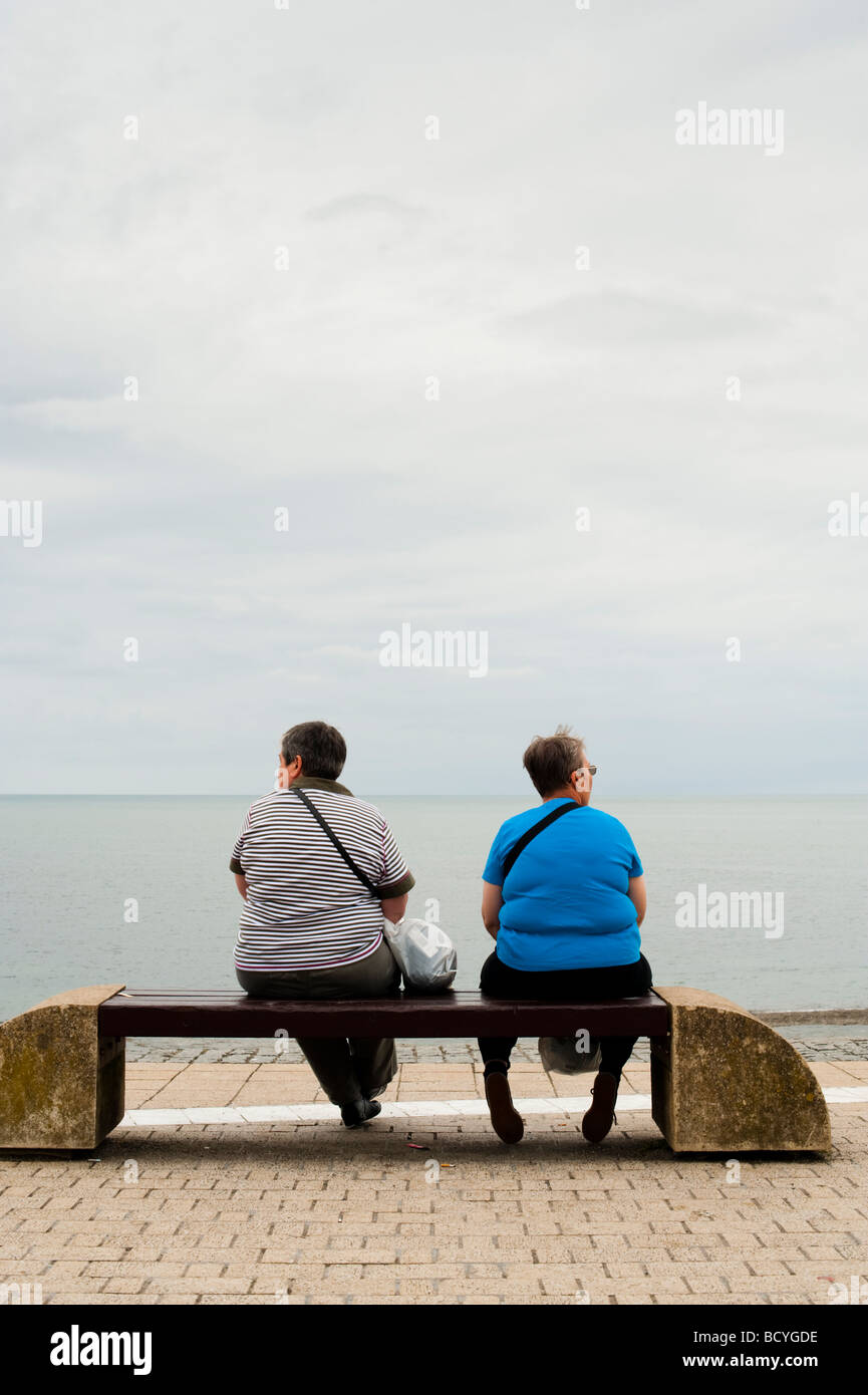 Rückansicht der beiden übergewichtigen Frauen im mittleren Alter sitzen auf einer Bank am Meer auf einem bewölkten grauen Sommer Tag UK Stockfoto