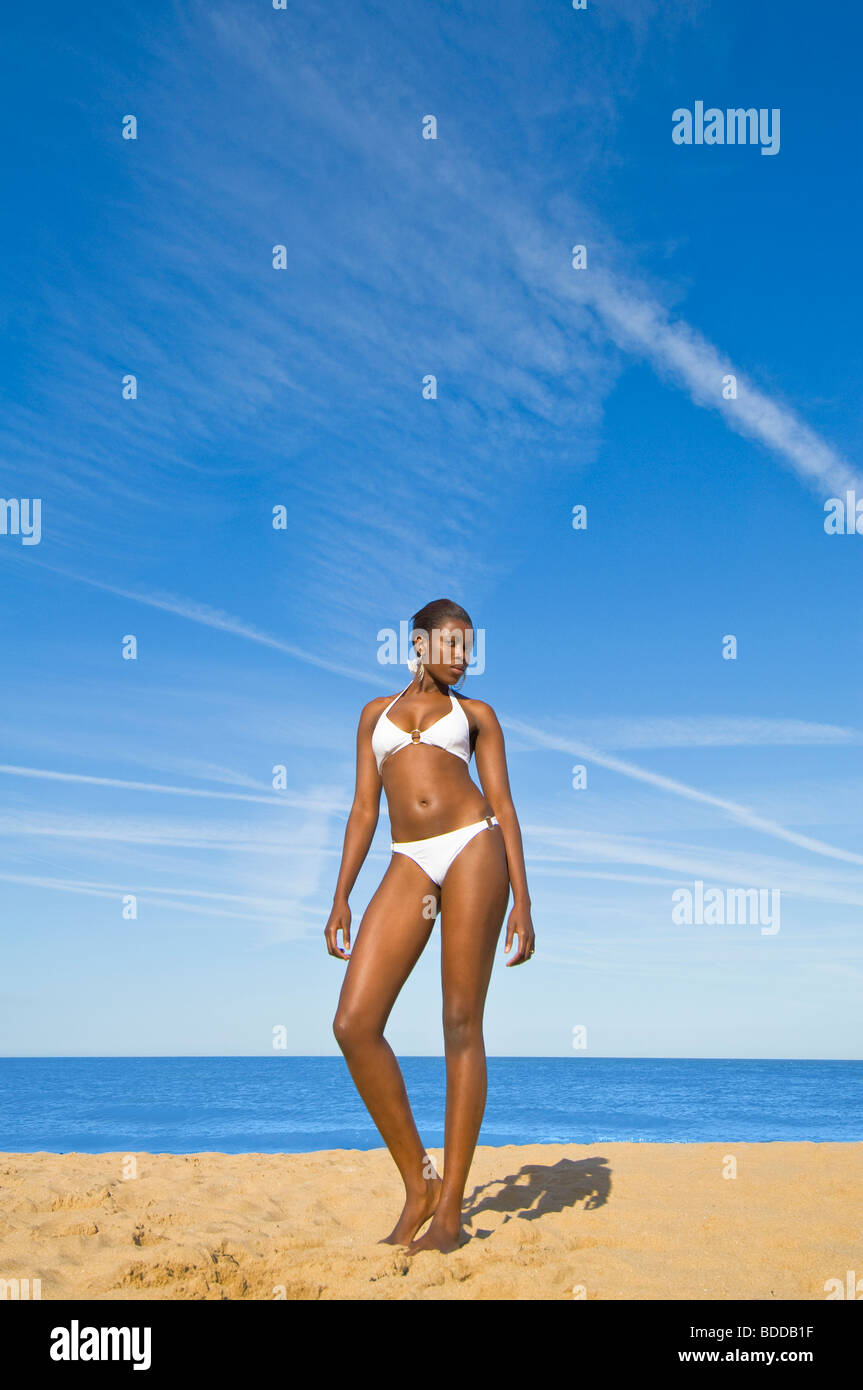 Naheinstellung Weitwinkel von eine schlanke attraktive afrikanische Frau posiert auf einem sandigen Strand mit einem blauen Meer und Himmel im Hintergrund. Stockfoto