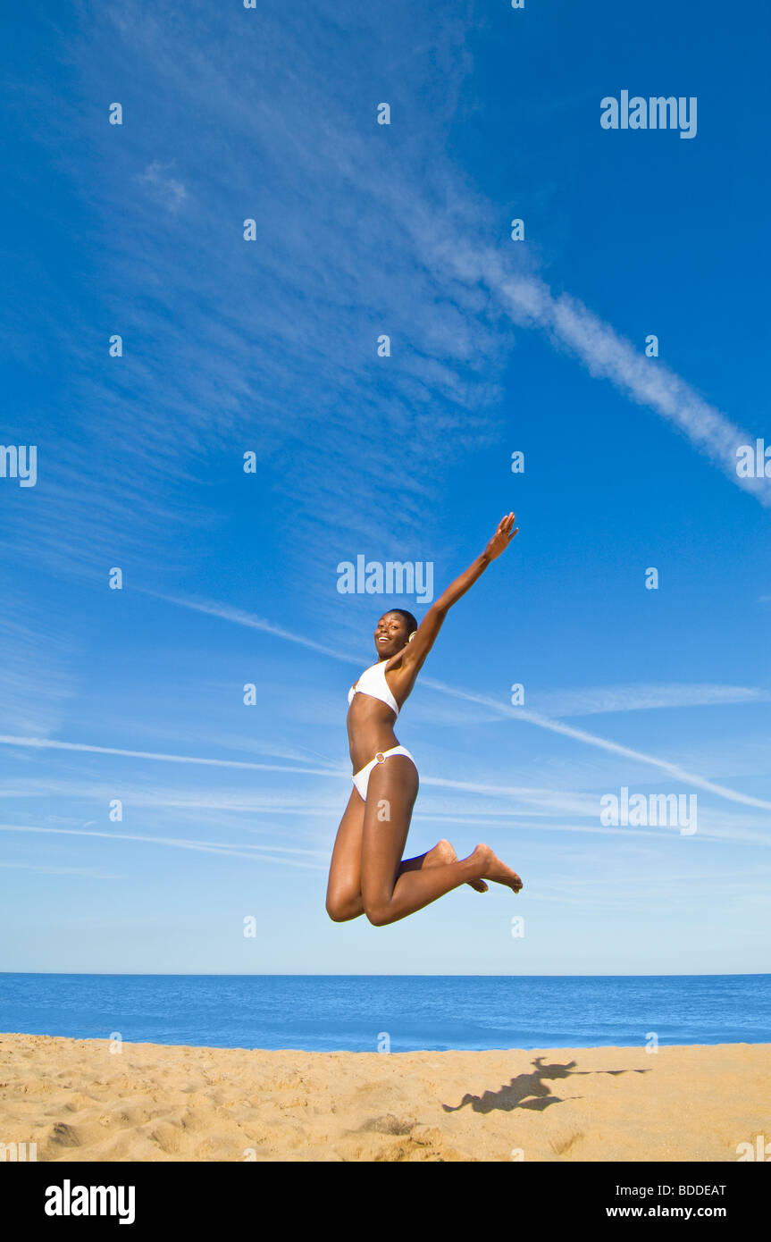 Naheinstellung Weitwinkel von eine schlanke attraktive Afrikanerin springen auf einem sandigen Strand mit einem blauen Meer und Himmel im Hintergrund. Stockfoto