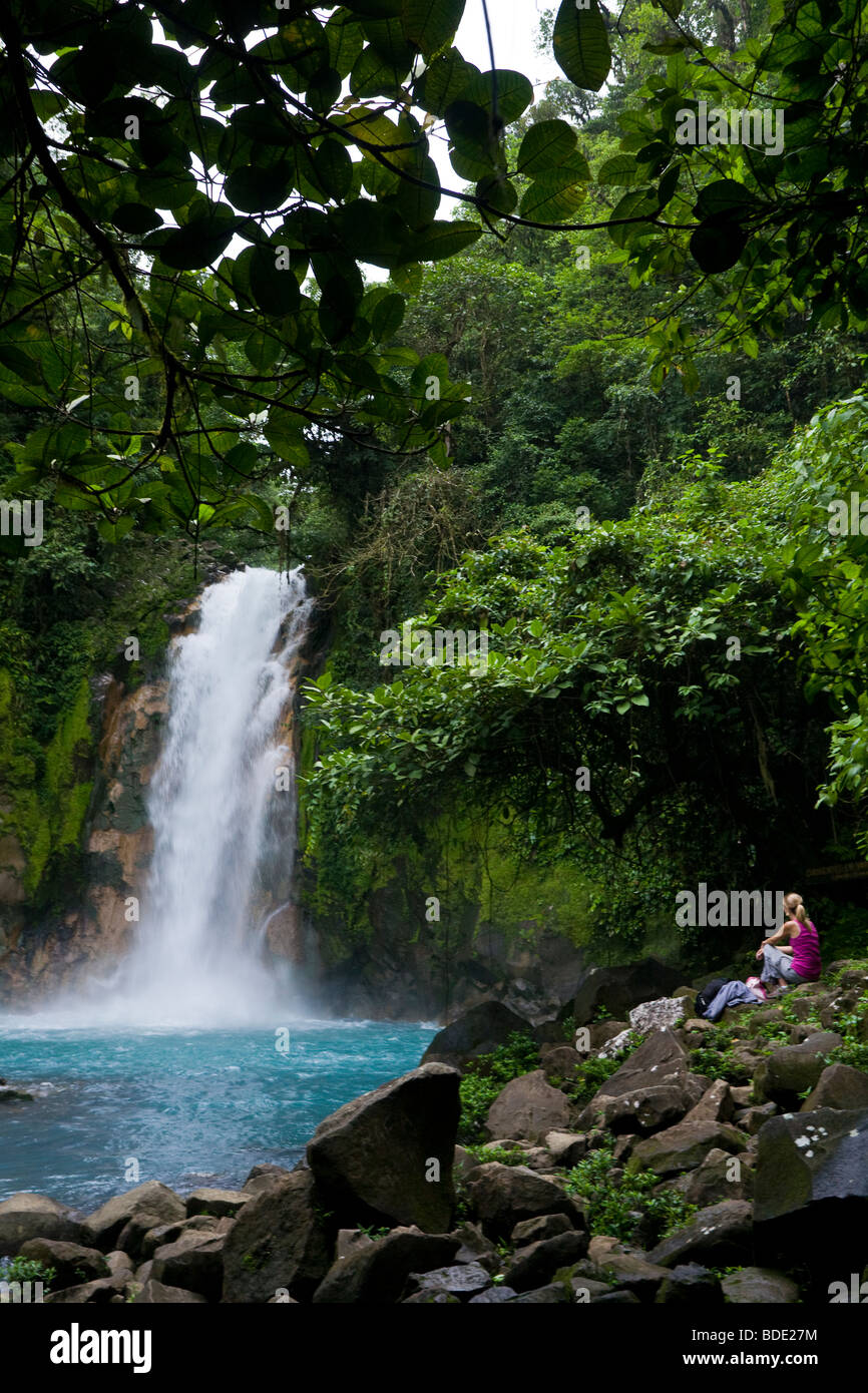 Weibliche Wanderer ruht unter einem Wasserfall entlang des pulsierenden blauen Rio Celeste Flusses im Vulkan-Nationalpark Tenorio, Costa Rica. Stockfoto